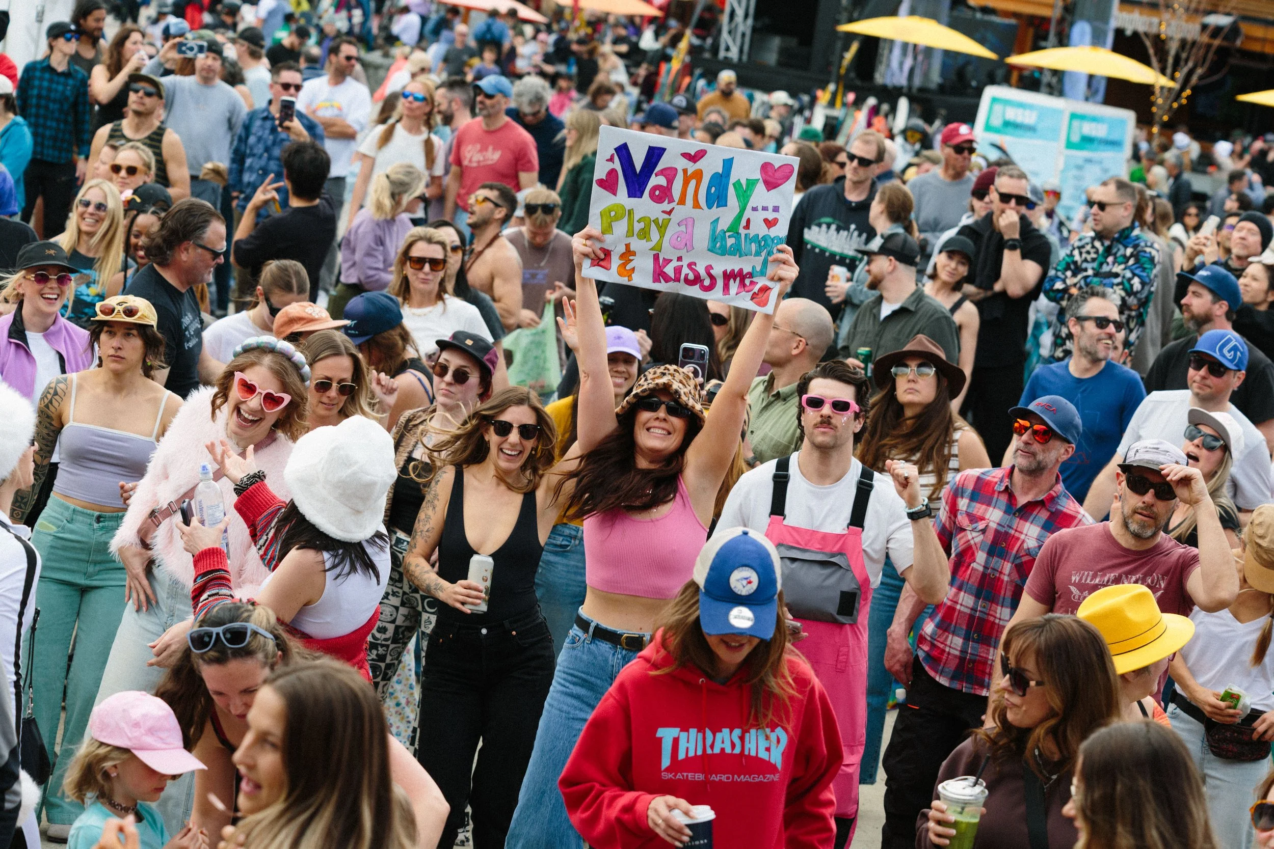 Crowd of people at an outdoor event, many smiling and wearing sunglasses, with a woman holding a colorful sign that says 'Vandy play a banner & kiss me'.