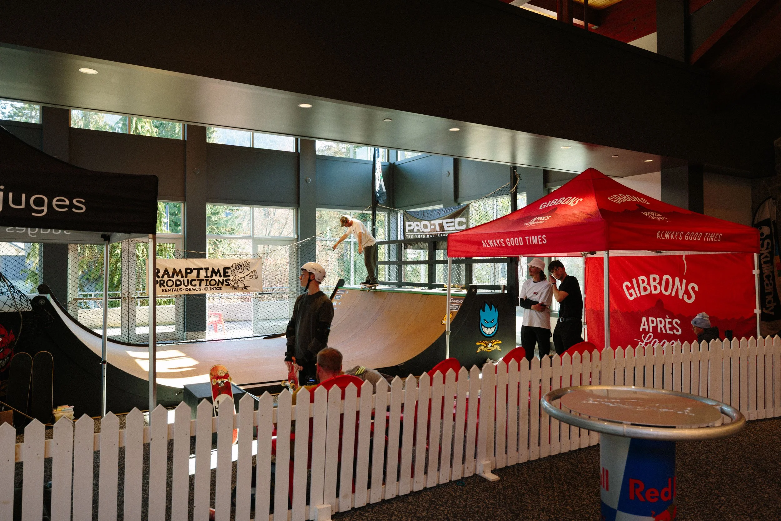 Indoor skatepark with a mini ramp, banners, and tents promoting skateboarding brands. People are skateboarding and observing.