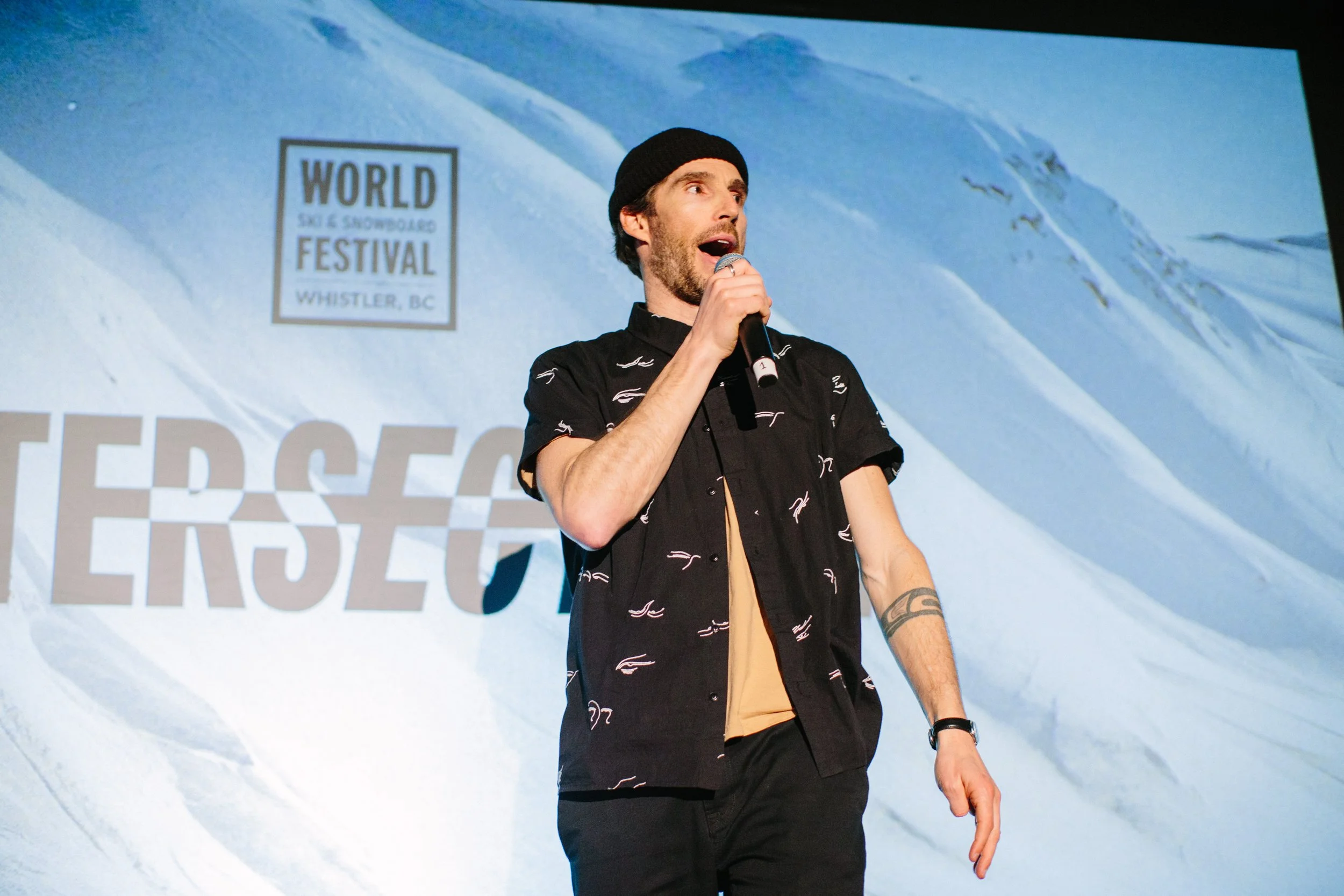 A man holding a microphone and speaking on stage at a snowboarding festival in Whistler, BC, with a backdrop of snow-covered mountains.