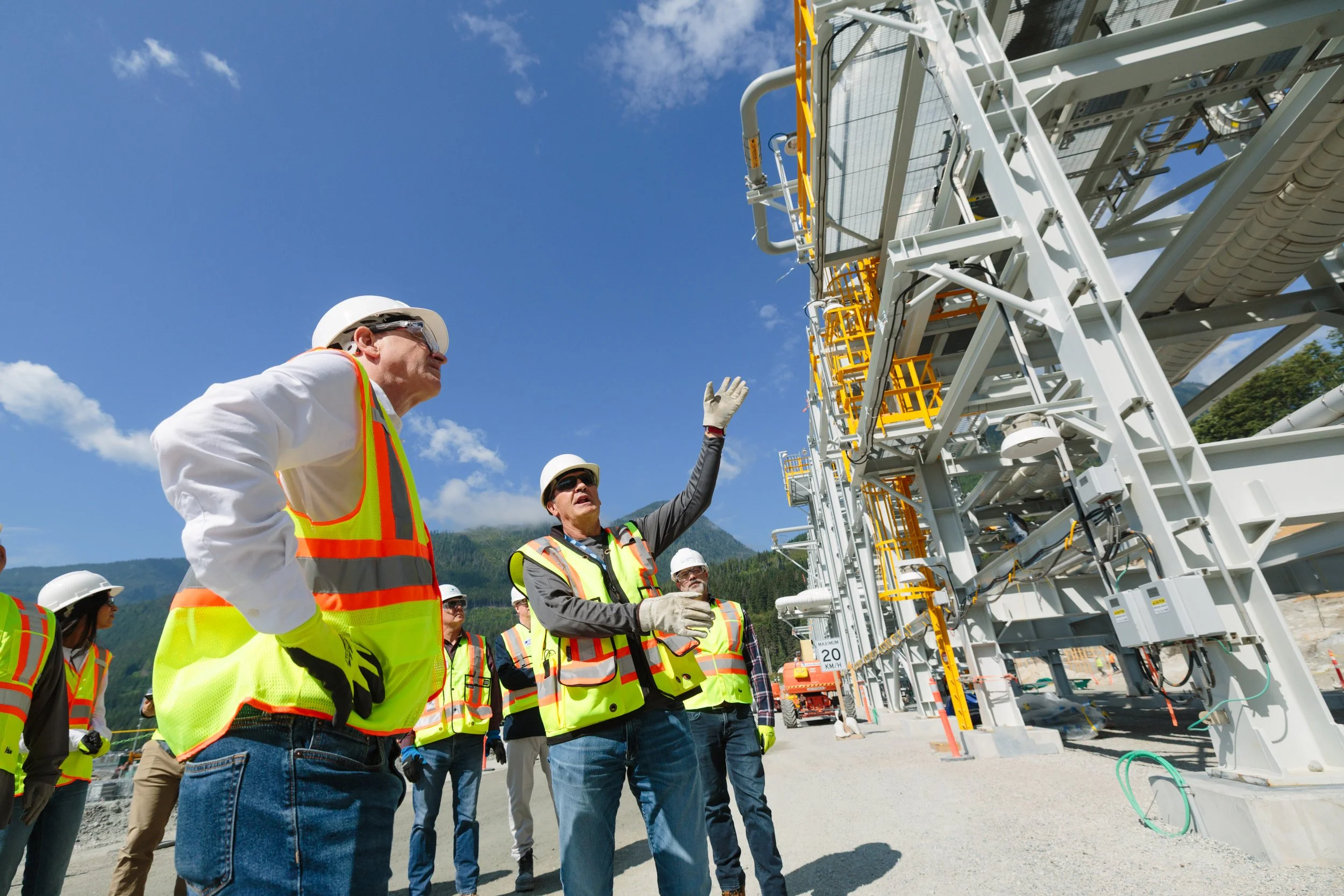 Group of construction workers wearing safety vests and helmets, gathered outdoors under a blue sky, inspecting a large industrial structure or machinery.