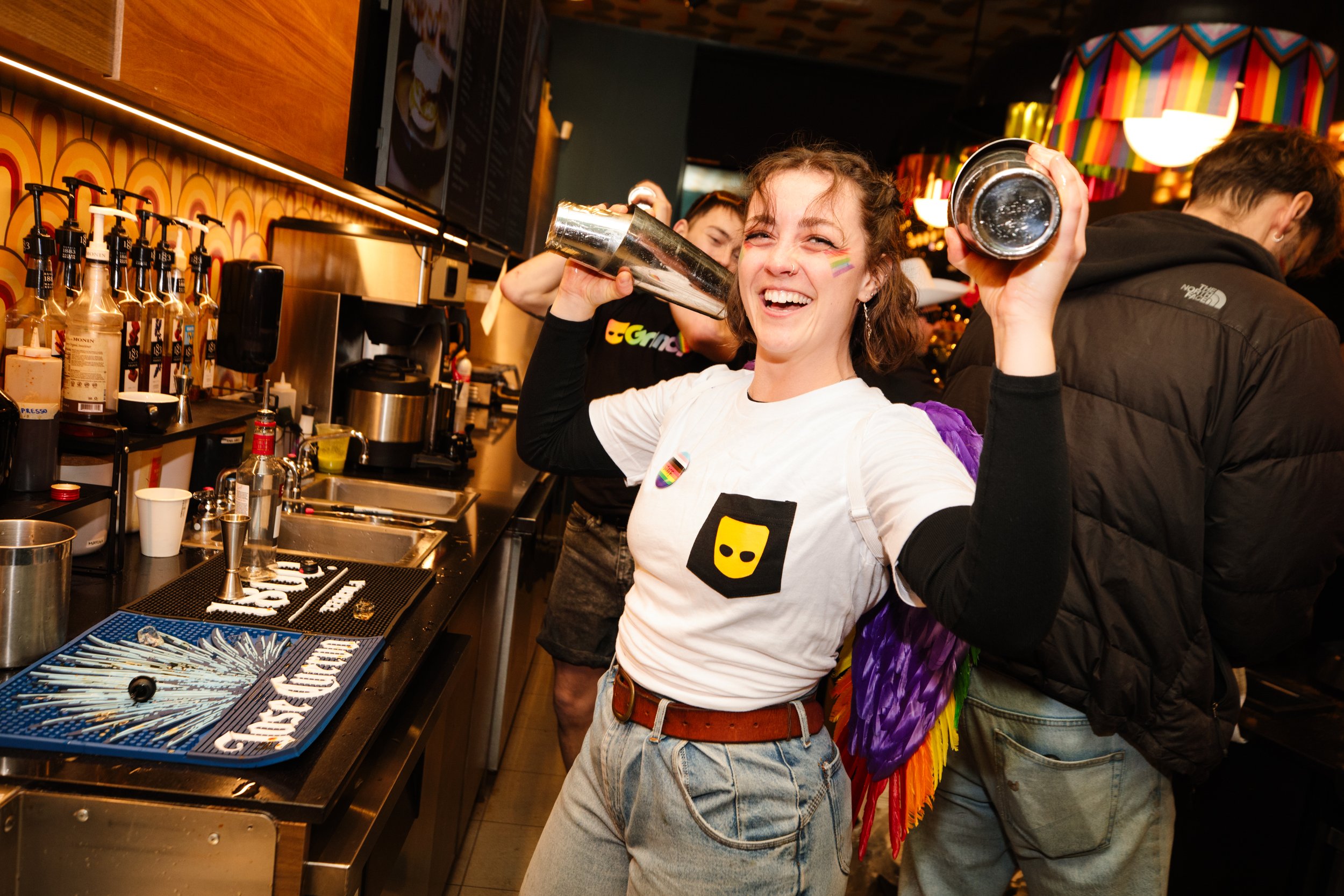 A smiling woman wearing a white T-shirt with patch and rainbow sticker, holding a cocktail shaker, at a bar with liquor bottles and mixers, surrounded by other people in a festive, colorful setting.