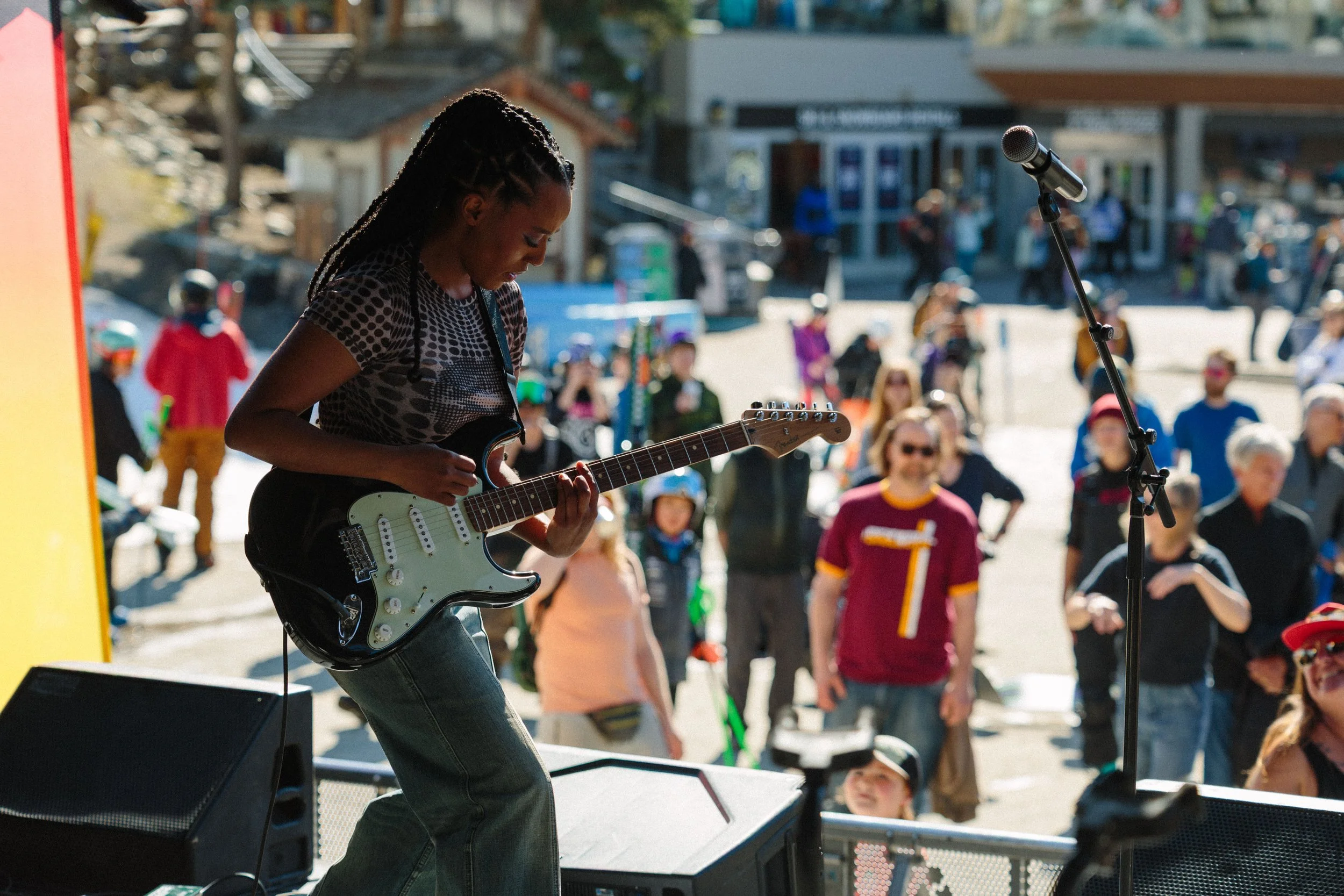 A woman playing an electric guitar on stage during an outdoor event with a crowd of people in the background.