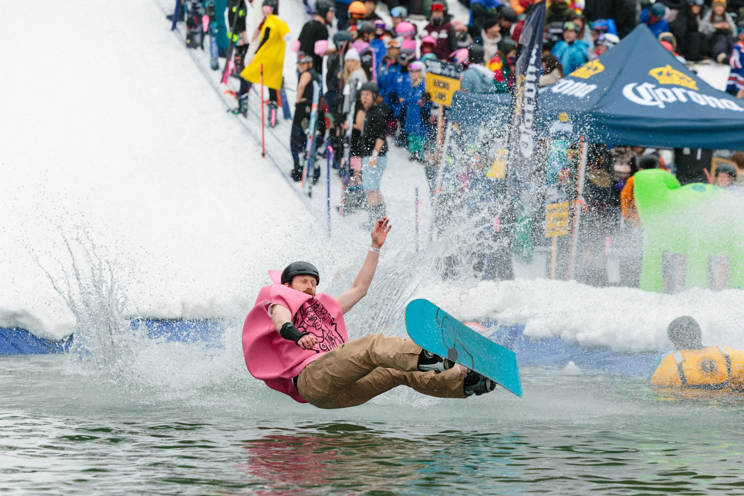 A person in a pink hoodie is falling off a skate or snowboard into a body of water during a winter event, with snow and a crowd of spectators and participants in the background.