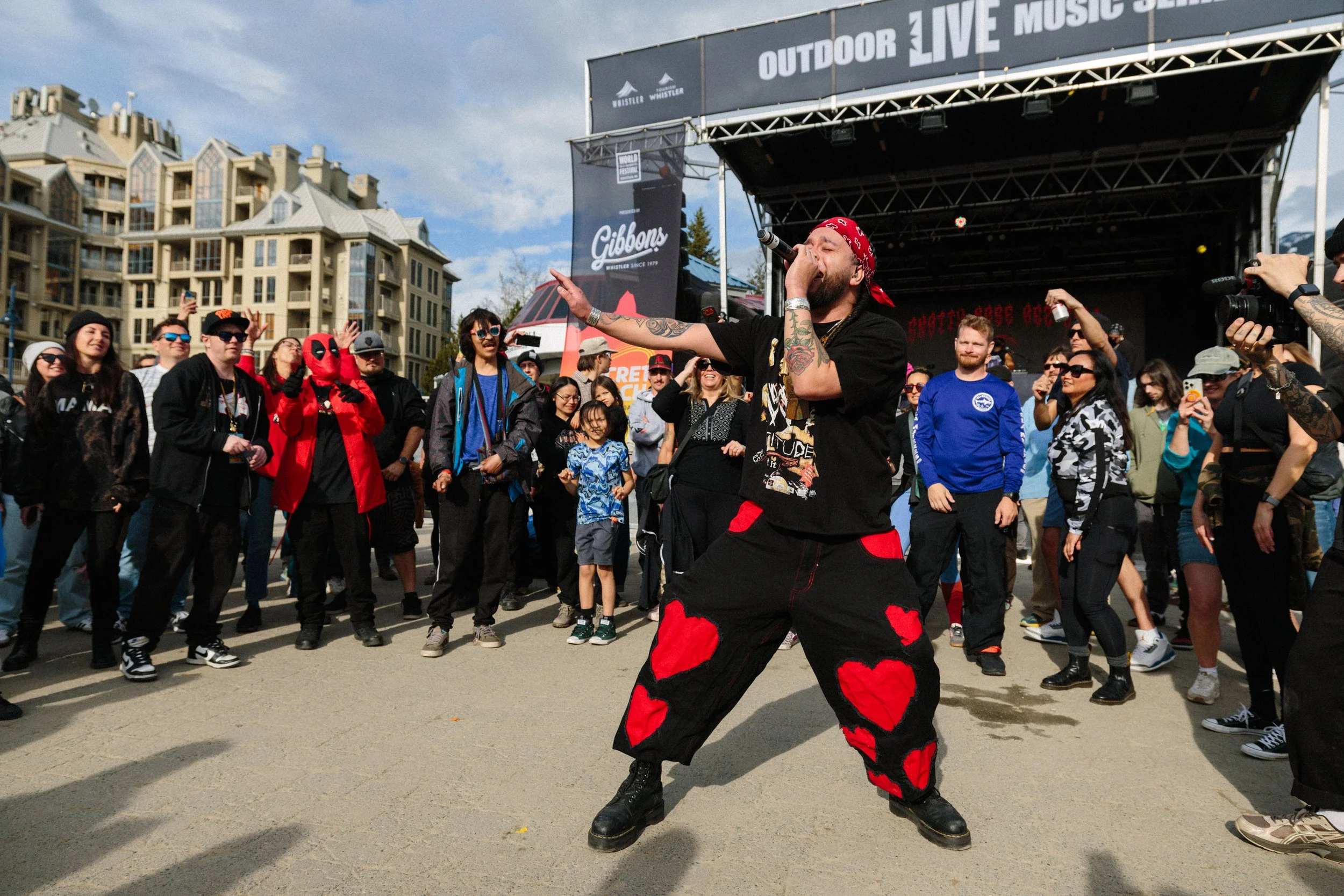 A performer with tattoos on their arm wearing a red bandana, black top, and loose black and red pants dancing or moving energetically in front of a crowd at an outdoor music event.