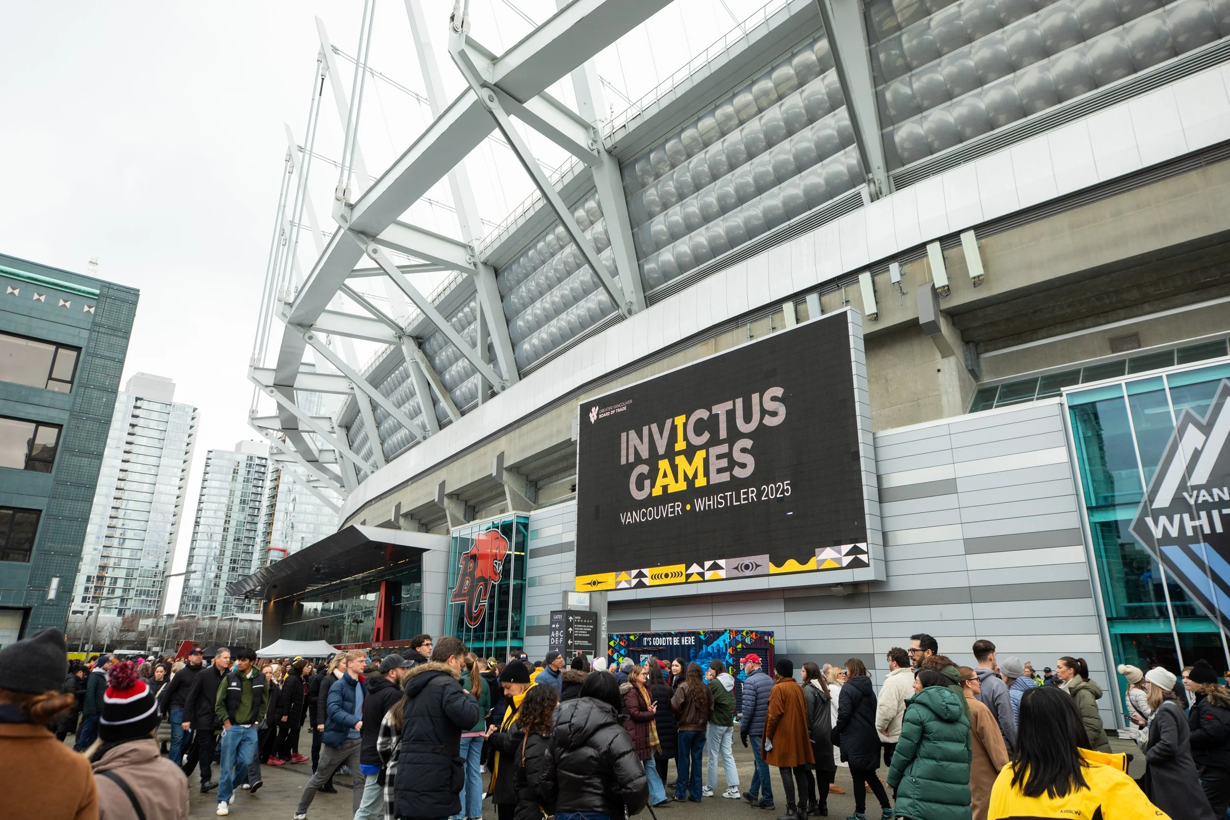 Crowd of people gathered outside a stadium for the Invictus Games Vancouver Whistler 2025, with a large digital screen displaying the event name and location.