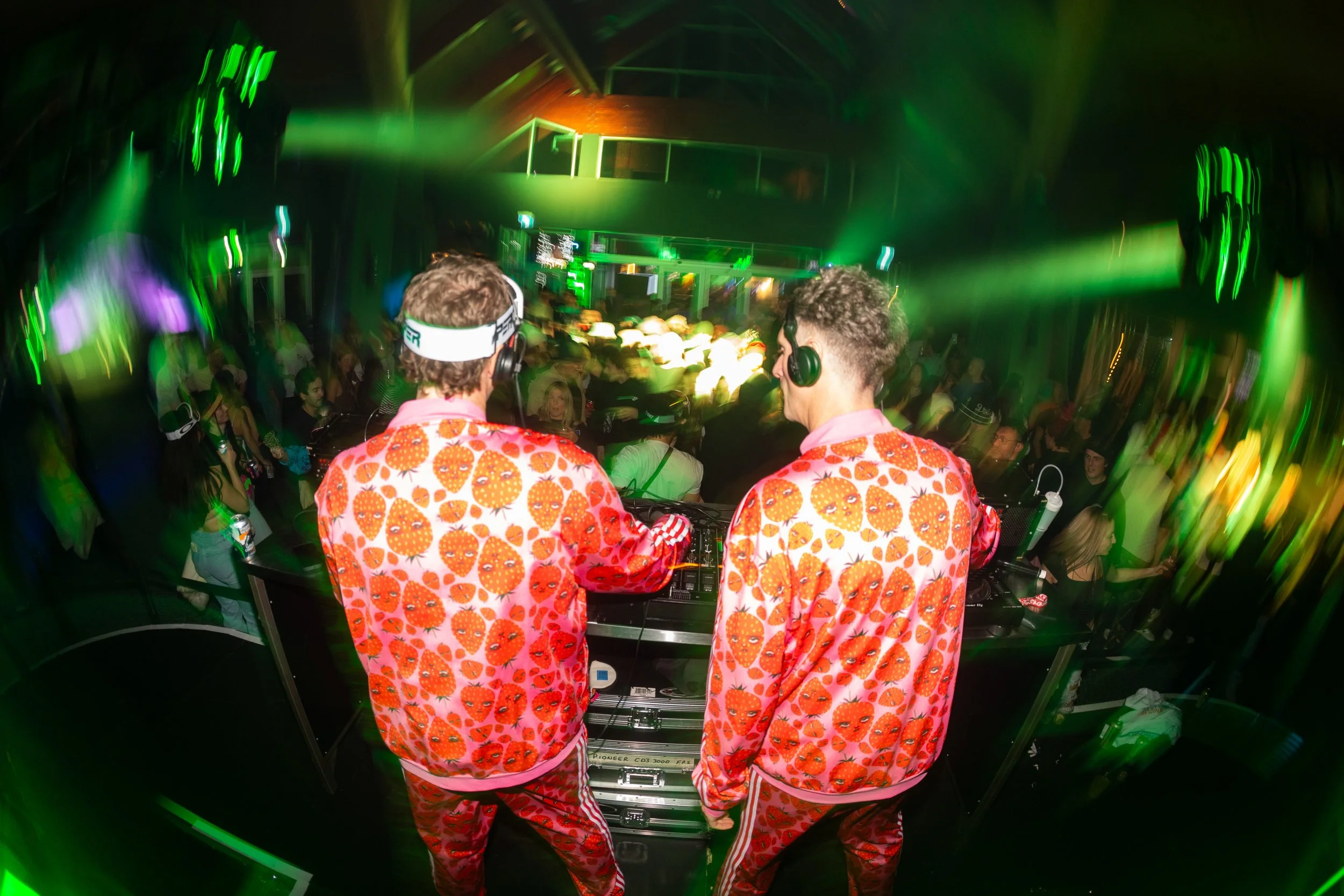 Two DJs in matching strawberry-patterned outfits playing music at a nightclub with green lighting and a dancing crowd.
