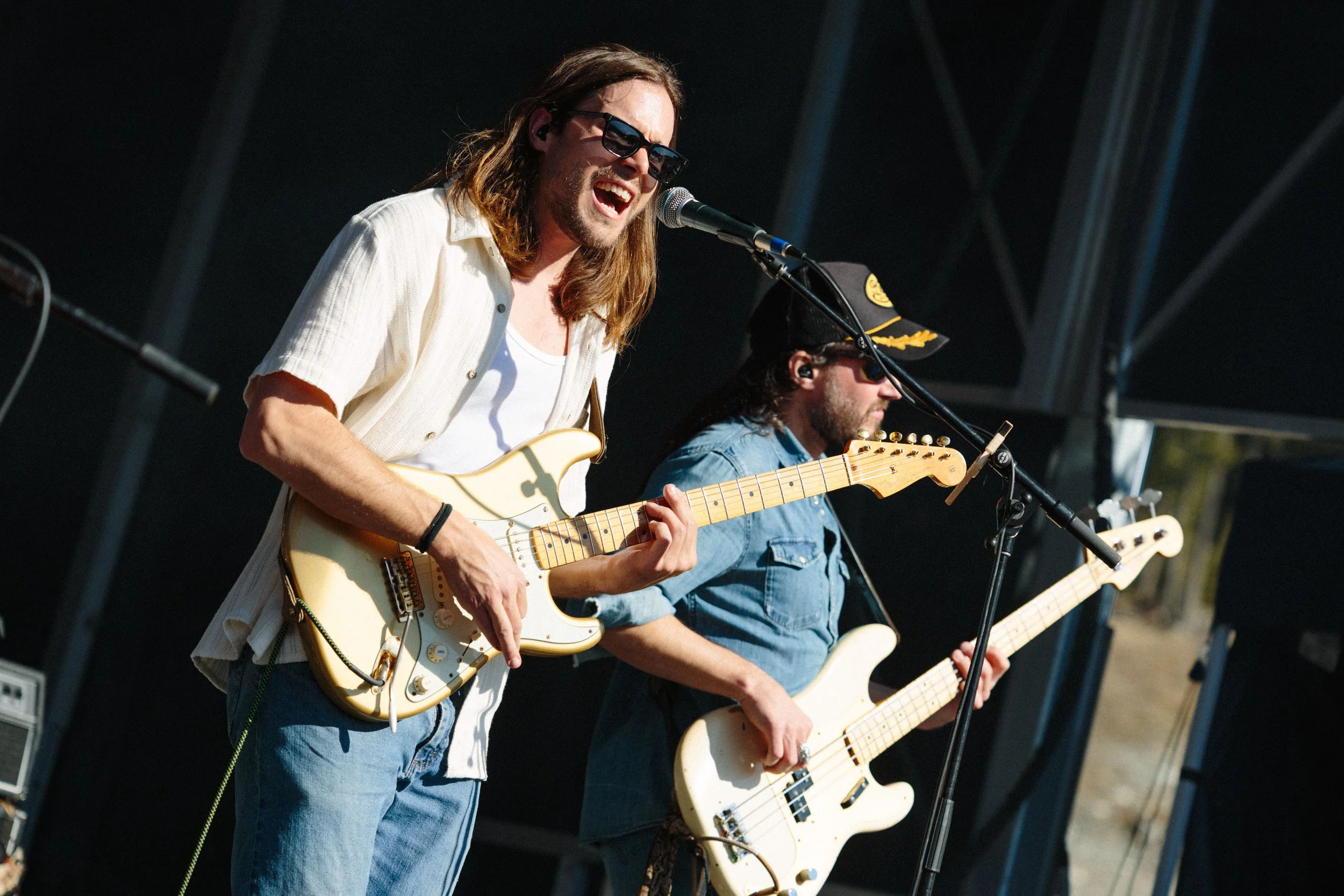 Two musicians playing electric guitars and singing into microphones on an outdoor stage during a performance.