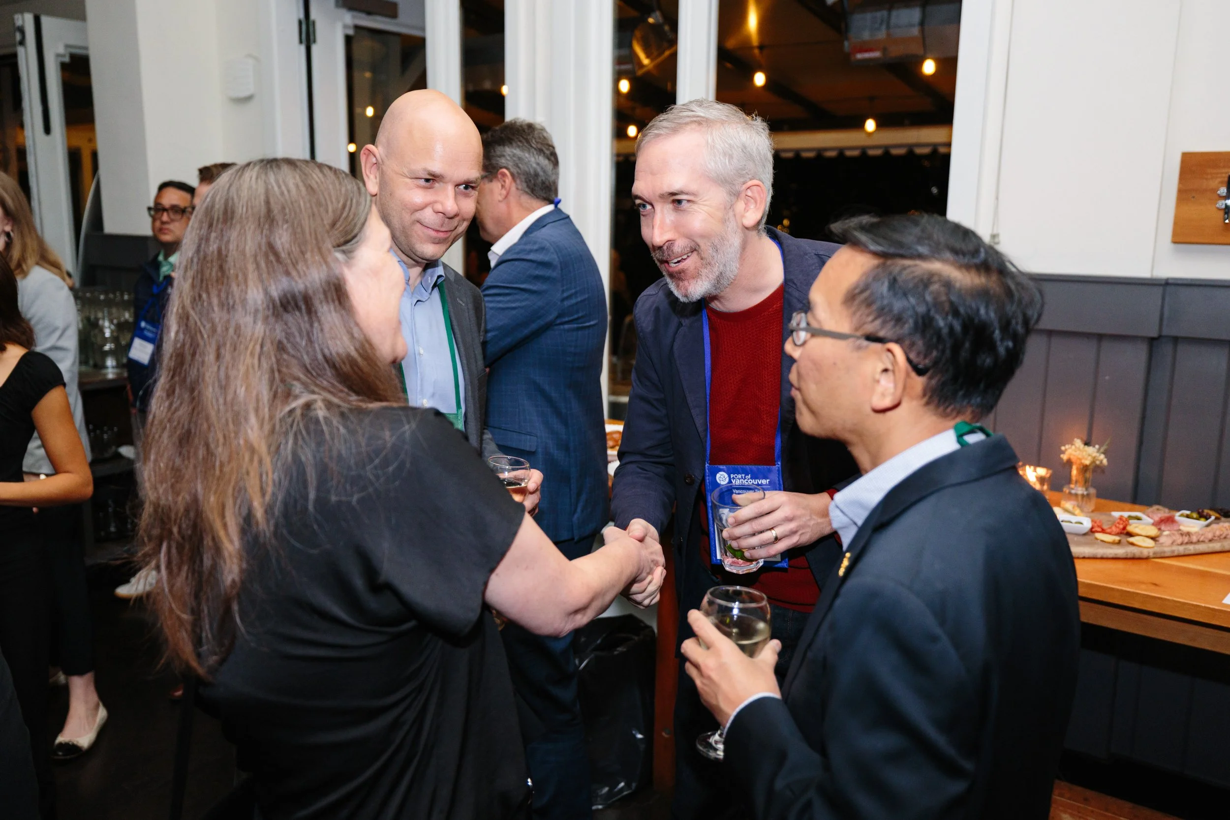 Group of five adults at a networking event, two men shaking hands, holding drinks, in a room with large windows and string lights, some people in the background near a table with snacks.
