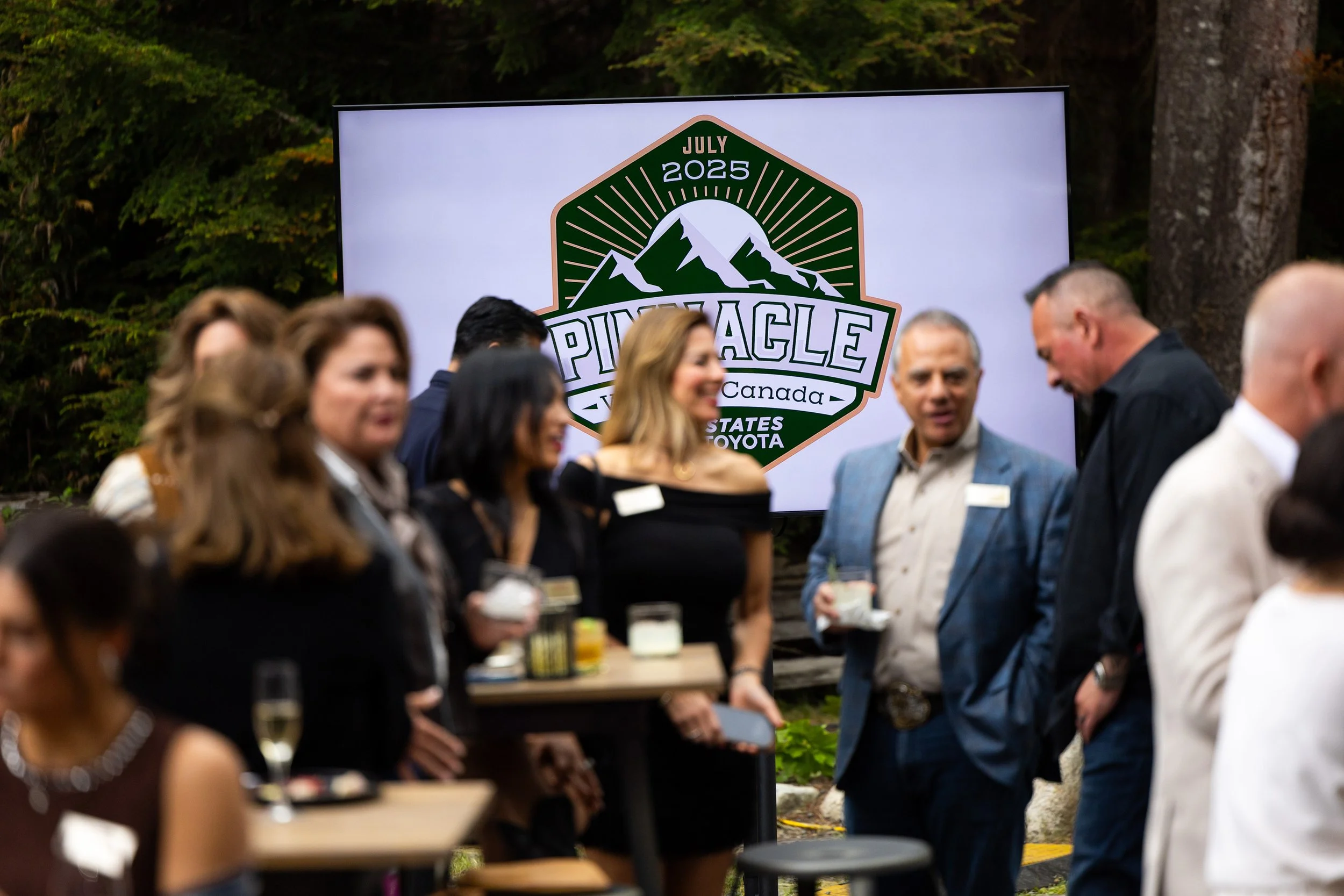 People attending an outdoor event with a large screen displaying a mountain and logo for Pinenacle Canada, July 2025.