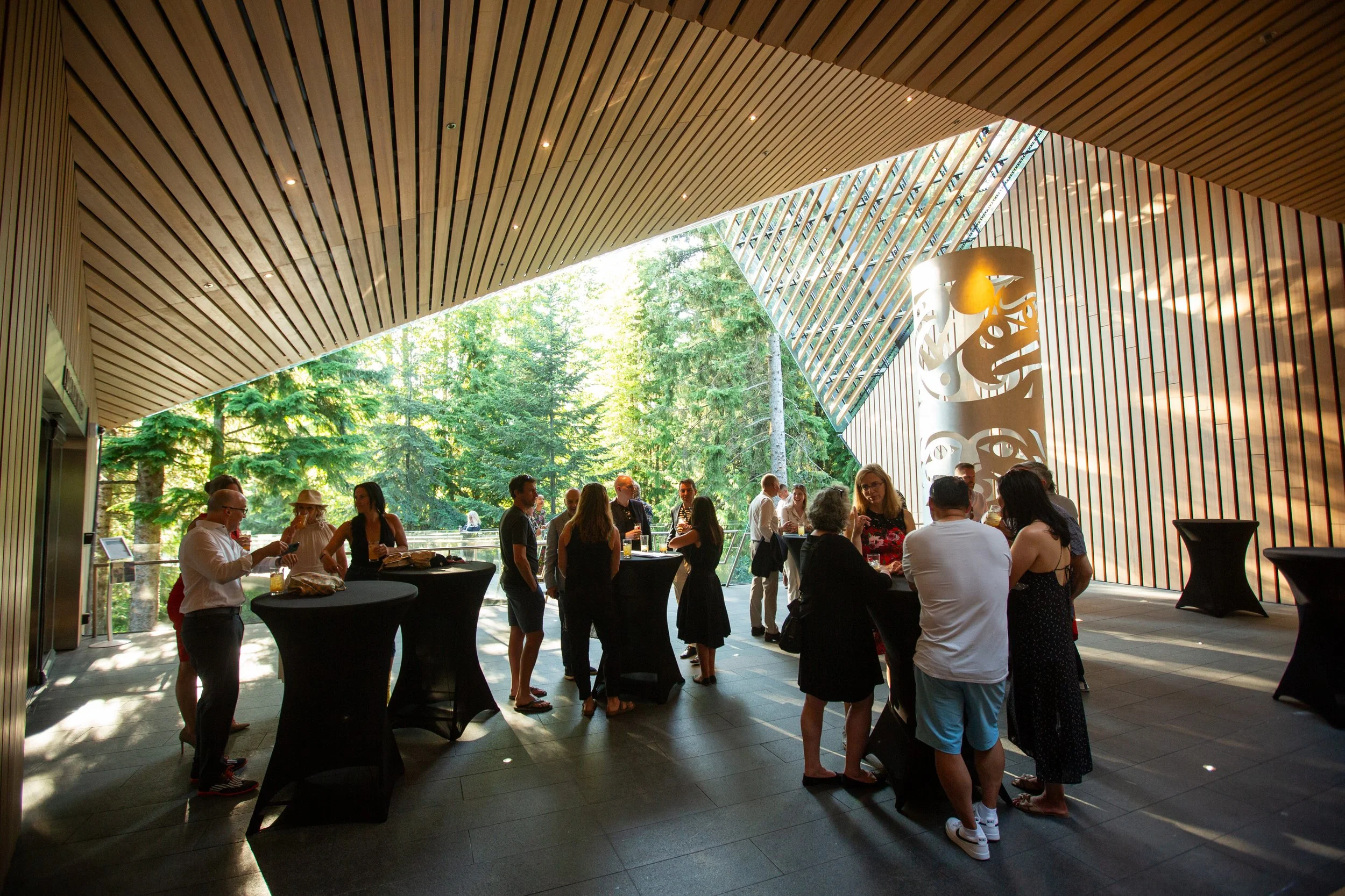 People socializing at a high-top cocktail event in a modern, open space with wood-paneled ceiling and large windows overlooking a forested area.