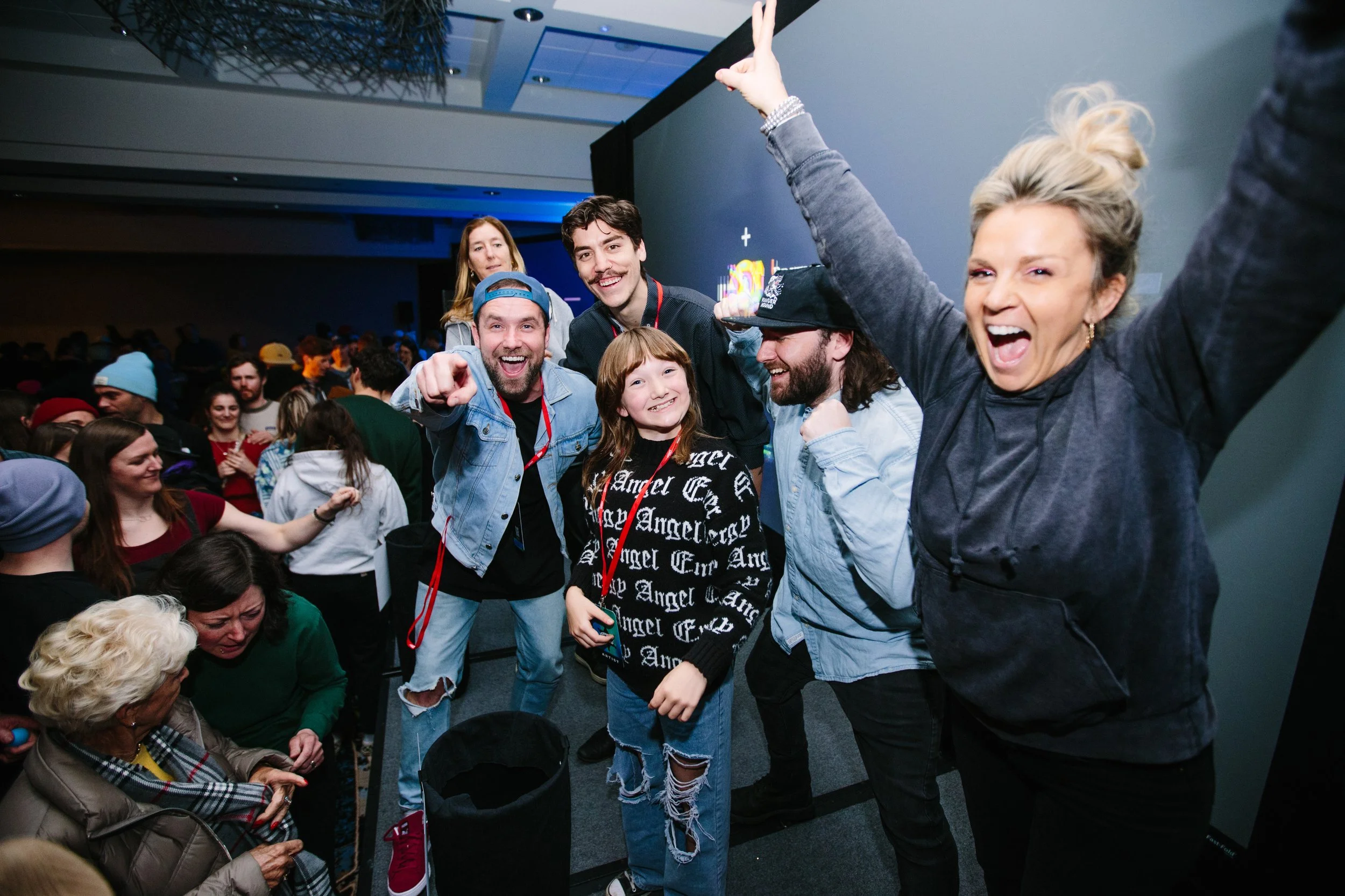 Group of young people and an older woman at an indoor event, smiling, taking a selfie, and making celebratory gestures. The environment appears festive with many attendees.
