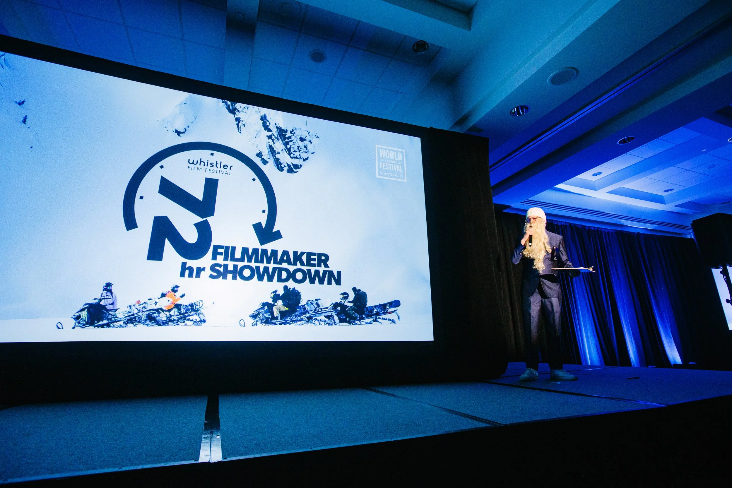 A person with a long white beard, wearing glasses, a white hat, a dark suit, and sneakers, standing on a stage and speaking into a microphone during a film festival event. There is a large screen behind him displaying a logo for the Whistler Film Fes