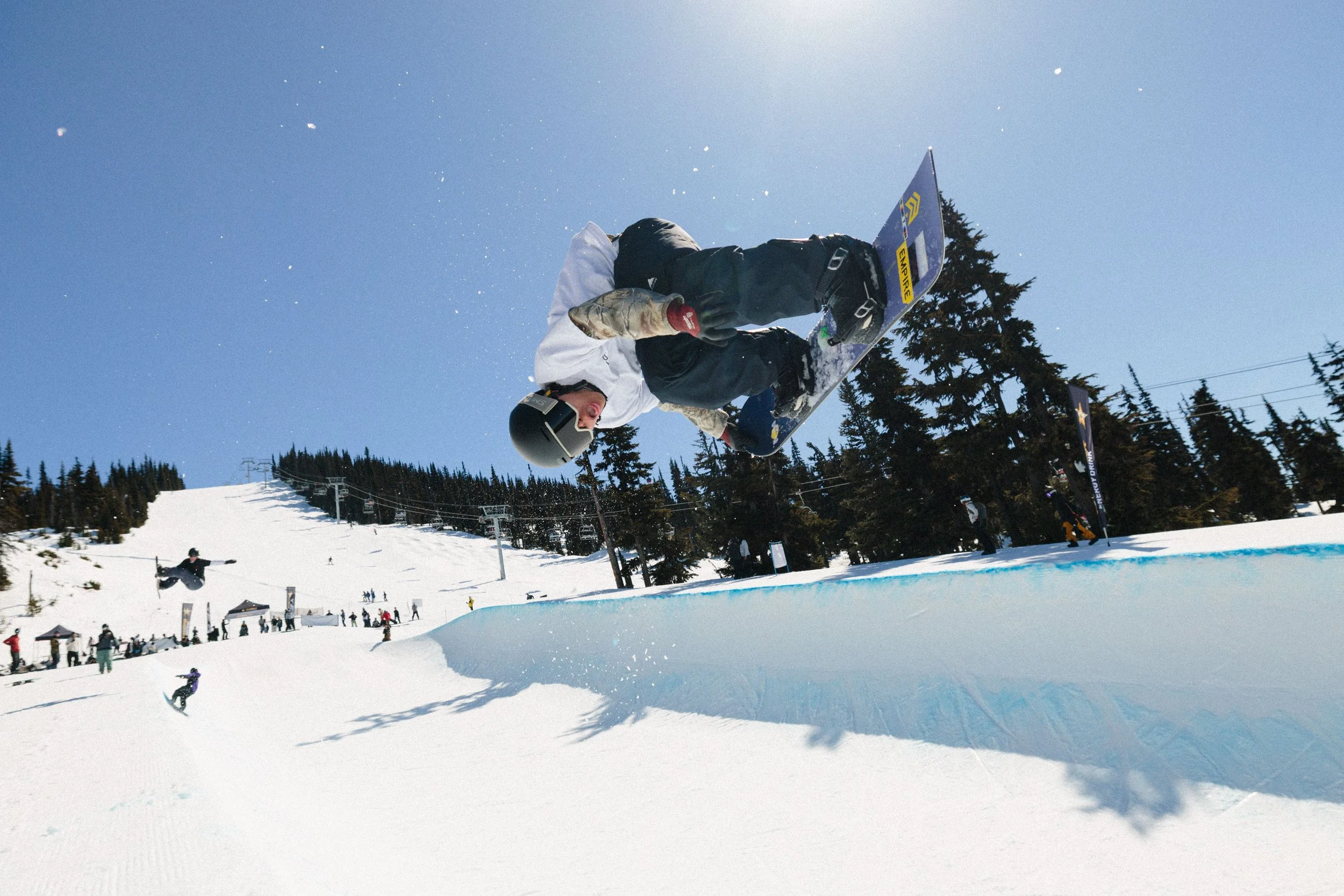 A person in winter sports gear performing a snowboard jump in a snowy ski resort with numerous spectators and other snowboarders on the hill, tall pine trees, and lift chairs in the background under a clear blue sky.