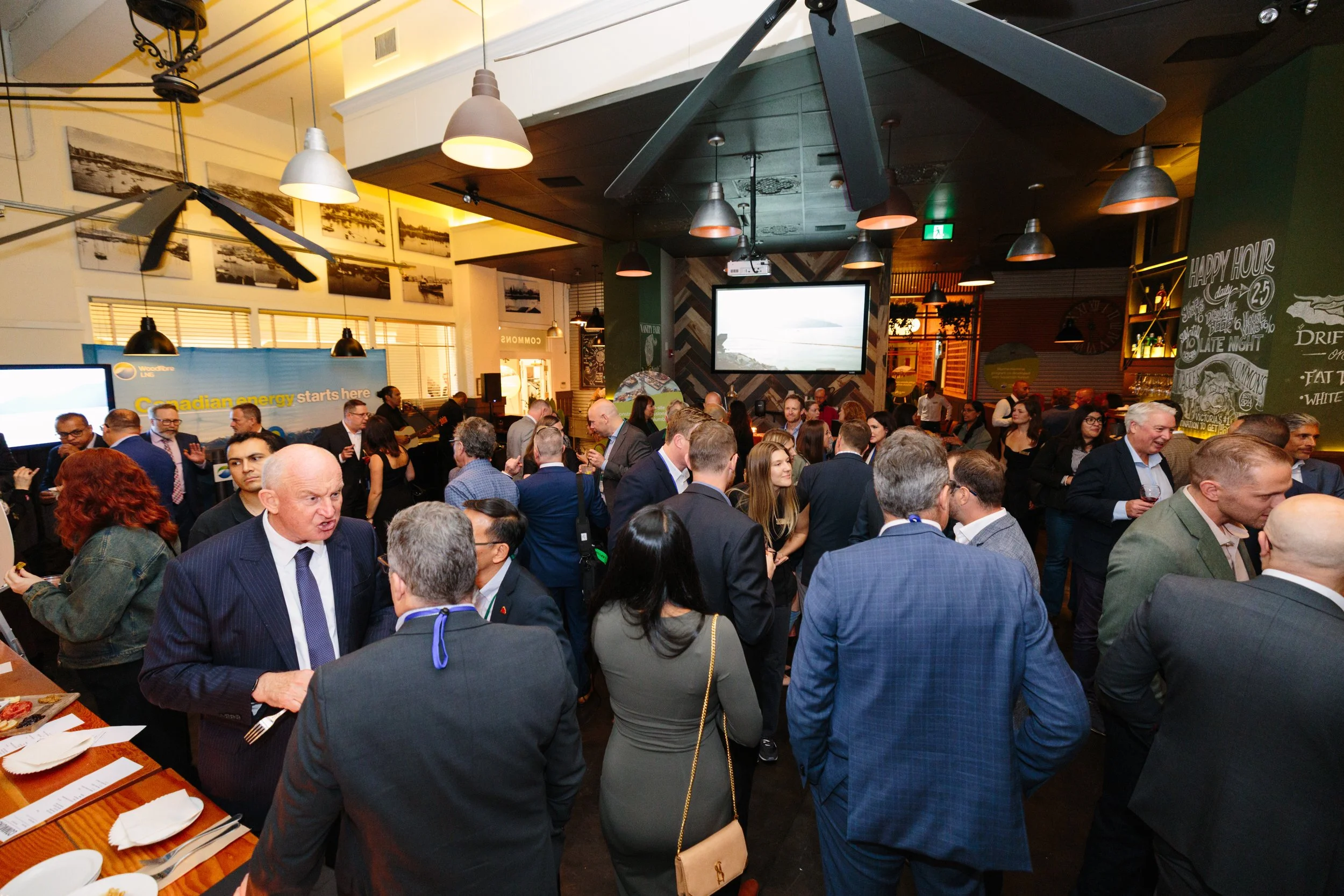 A crowded social event or business gathering in a restaurant or bar, with people dressed in business attire engaging in conversations and standing around tables, with a large screen showing a landscape and decorative wall chalkboard art in the backgr