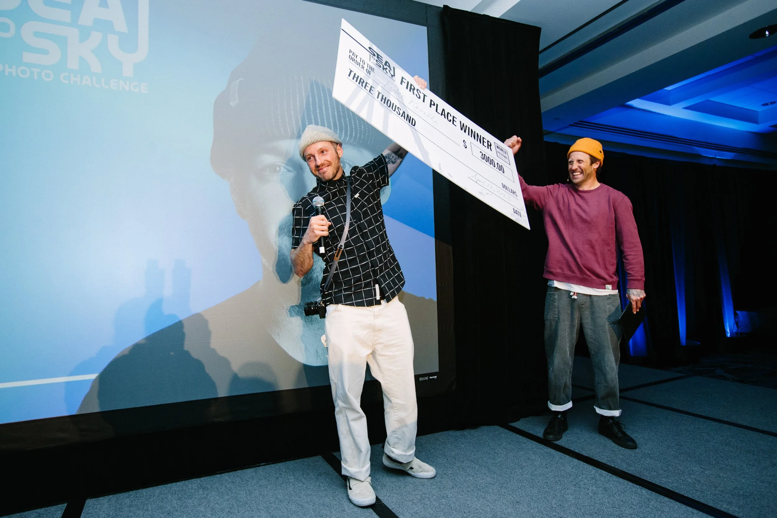 Two men on stage holding a large check for $3000, with one man speaking into a microphone. The check is for first place winner in a competition, and they are smiling, celebrating their victory.