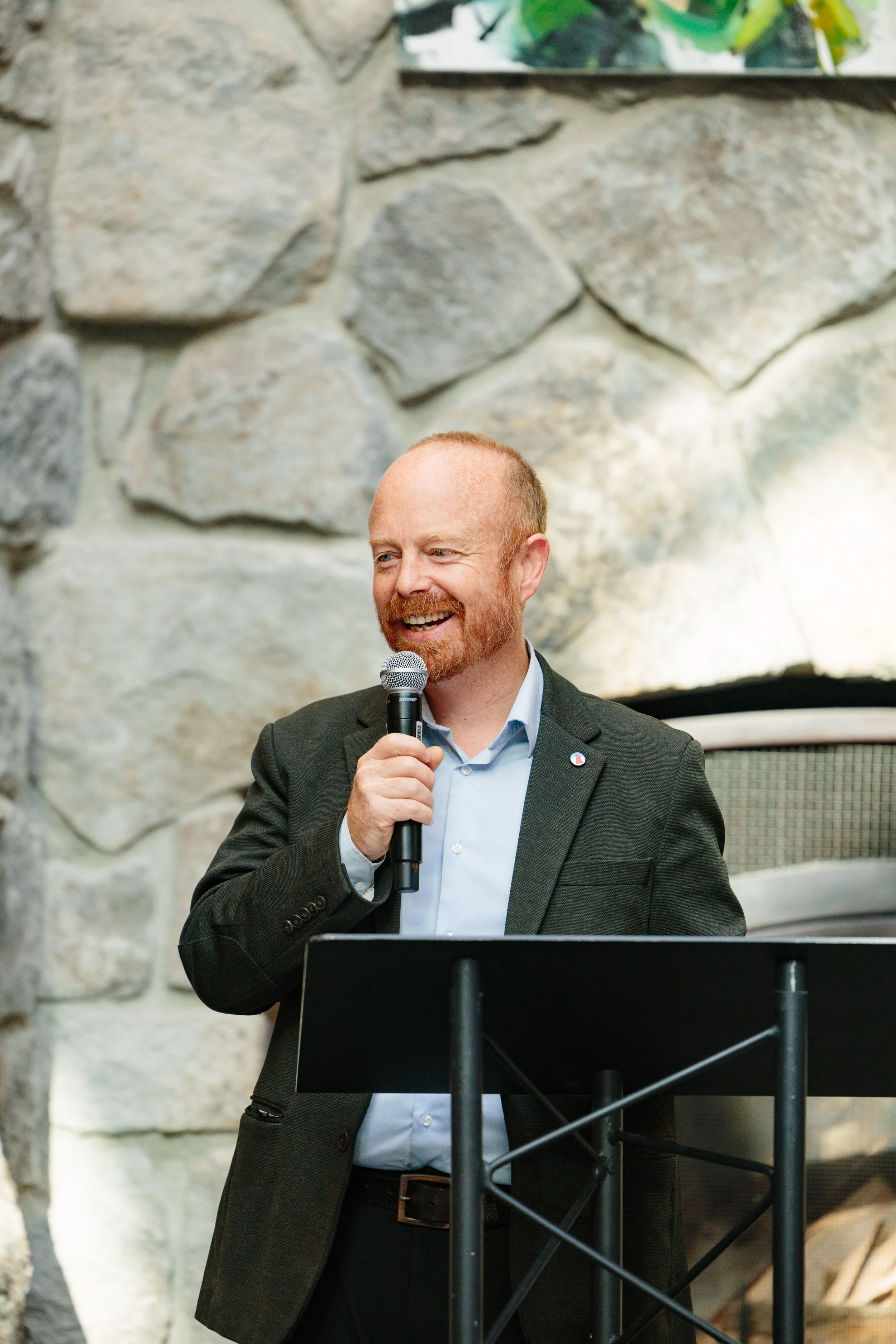 A man with red hair and a beard, wearing a dark blazer and a light blue shirt, is holding a microphone and speaking at a podium in front of a stone wall.