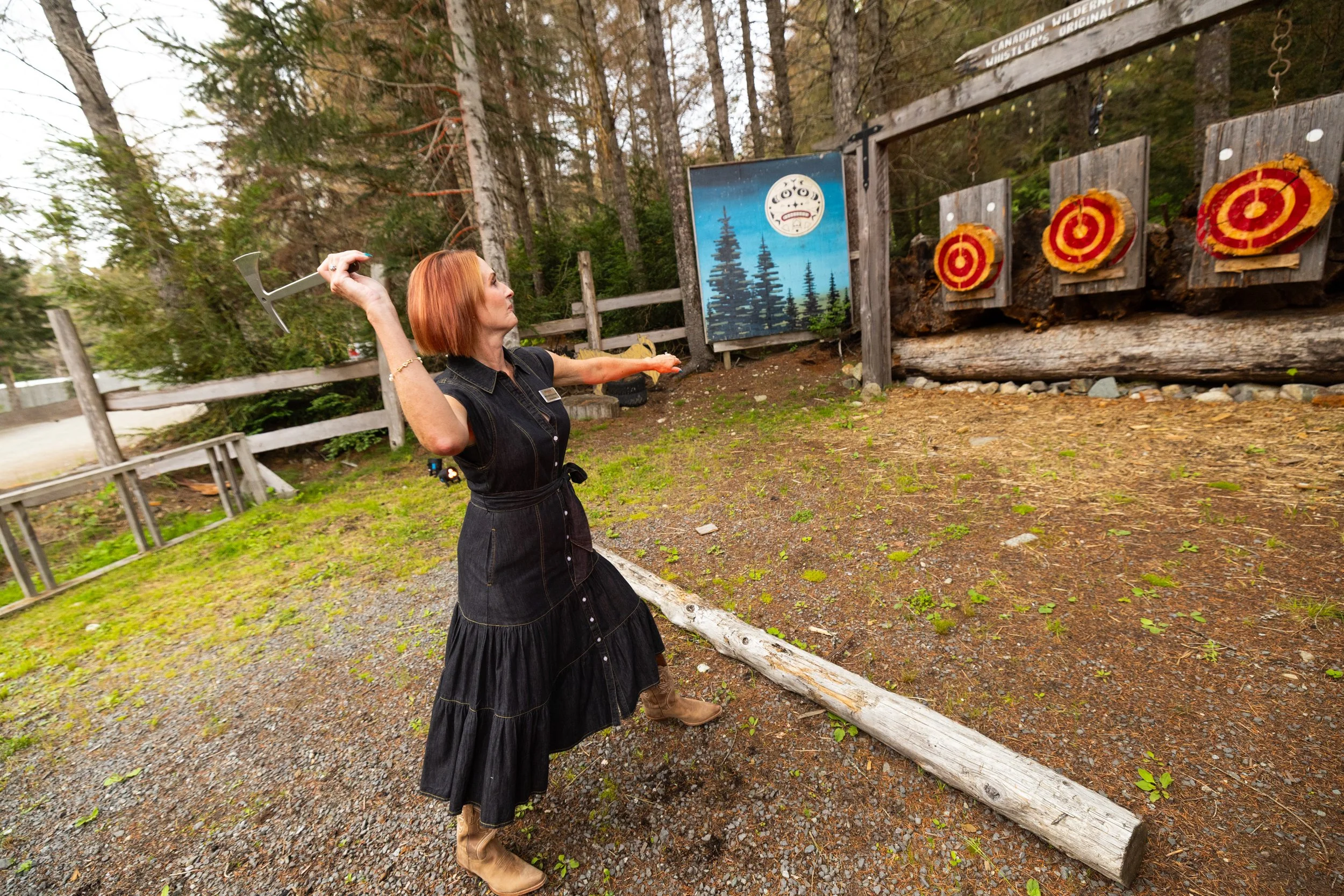 A woman with short red hair, wearing a black dress and tan boots, is playing axe throwing at an outdoor target range surrounded by trees. She is mid-throw with an axe in her right hand, aiming at three targets on a wooden panel. Behind her, there is 