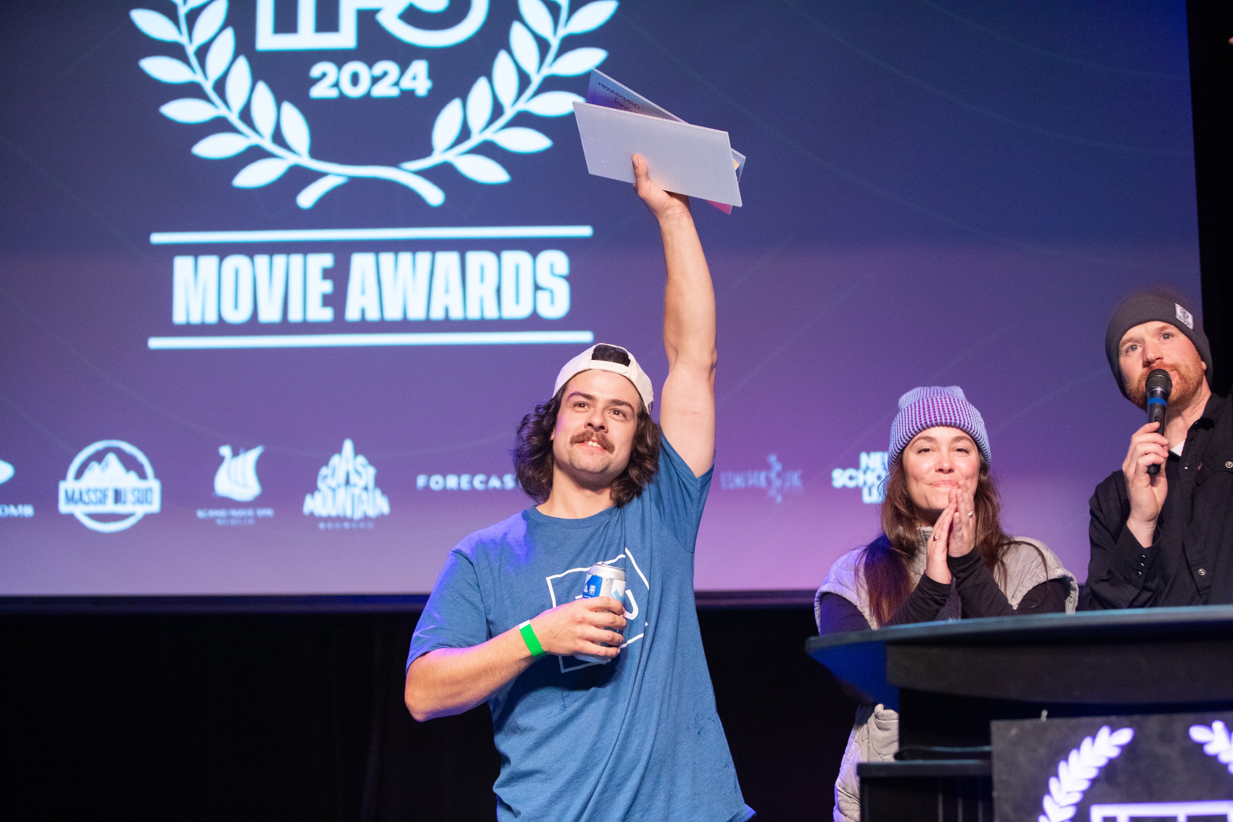 A man with long hair, mustache, wearing a baseball cap backward, holding a trophy above his head on stage at the 2024 Movie Awards. Two other people, a woman and a man, stand next to him. The woman claps and the man speaks into a microphone. A large 