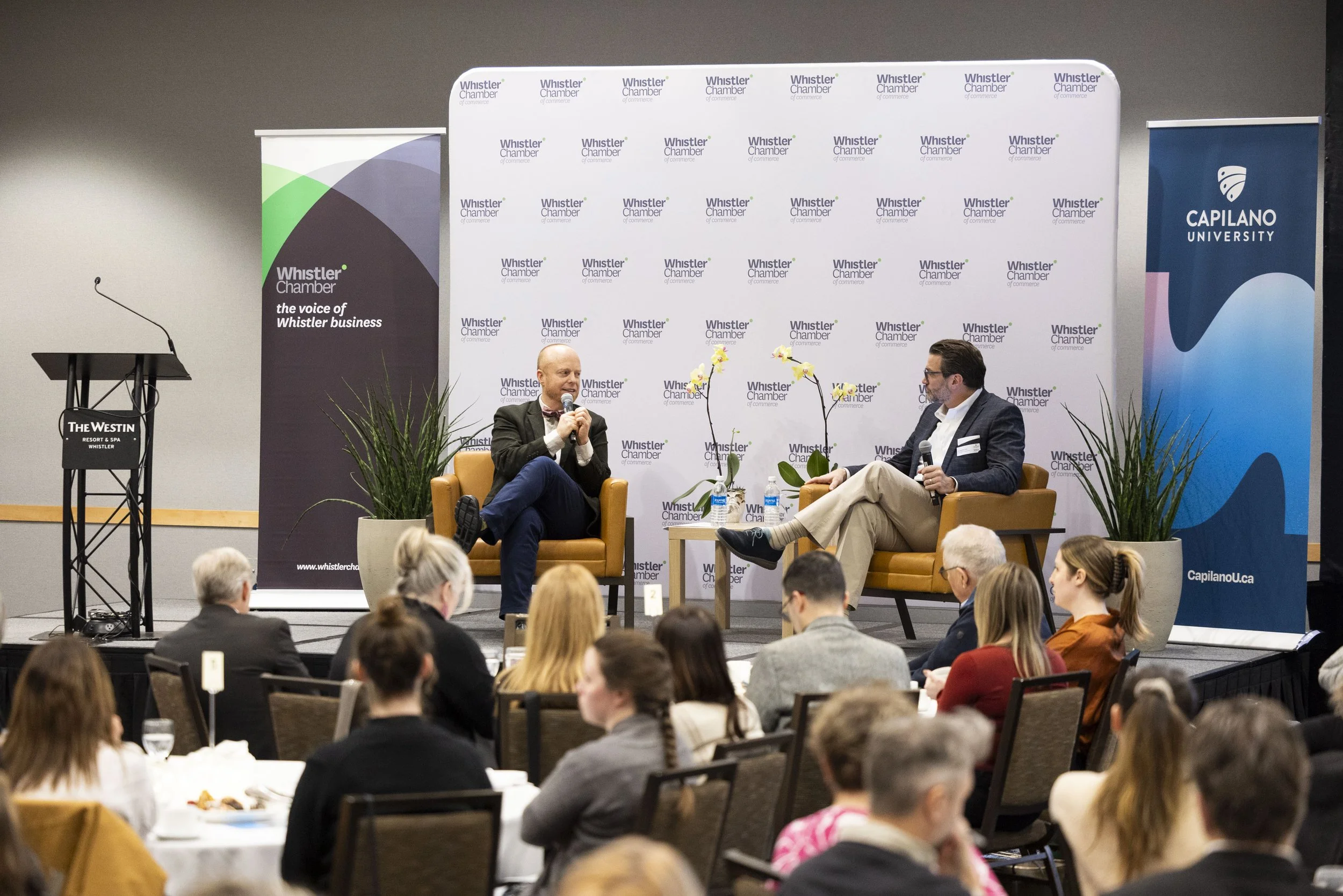 Two men are seated on stage in armchairs, engaging in a discussion with microphones, in front of an audience at a conference. Behind them are banners for Whistler Chamber and Capilano University.