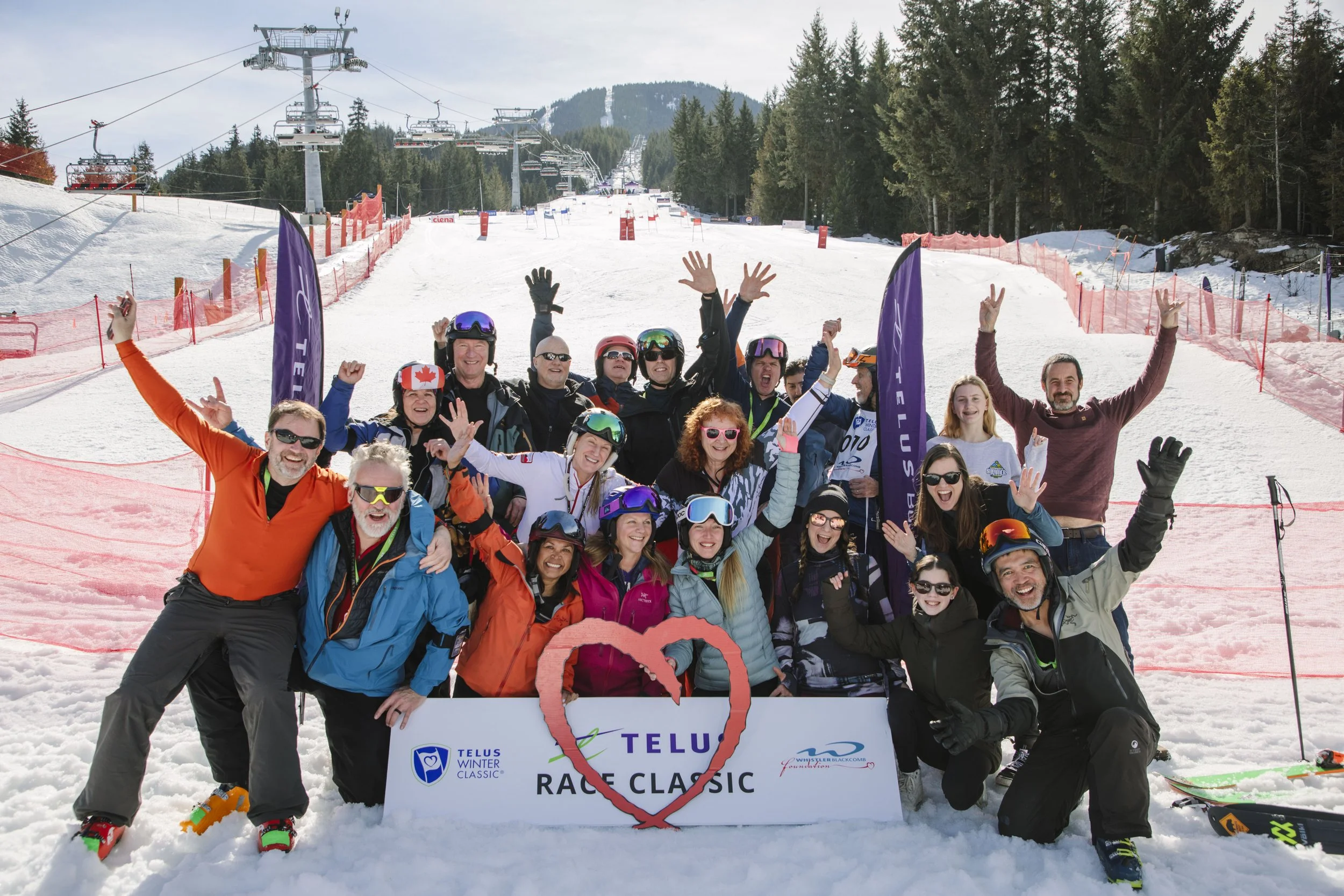 Group of skiers and snowboarders in colorful gear celebrating on a snowy mountain, holding a sign that reads 'TELUS Race Classic' with a red heart outline, ski lifts and evergreen trees in the background.