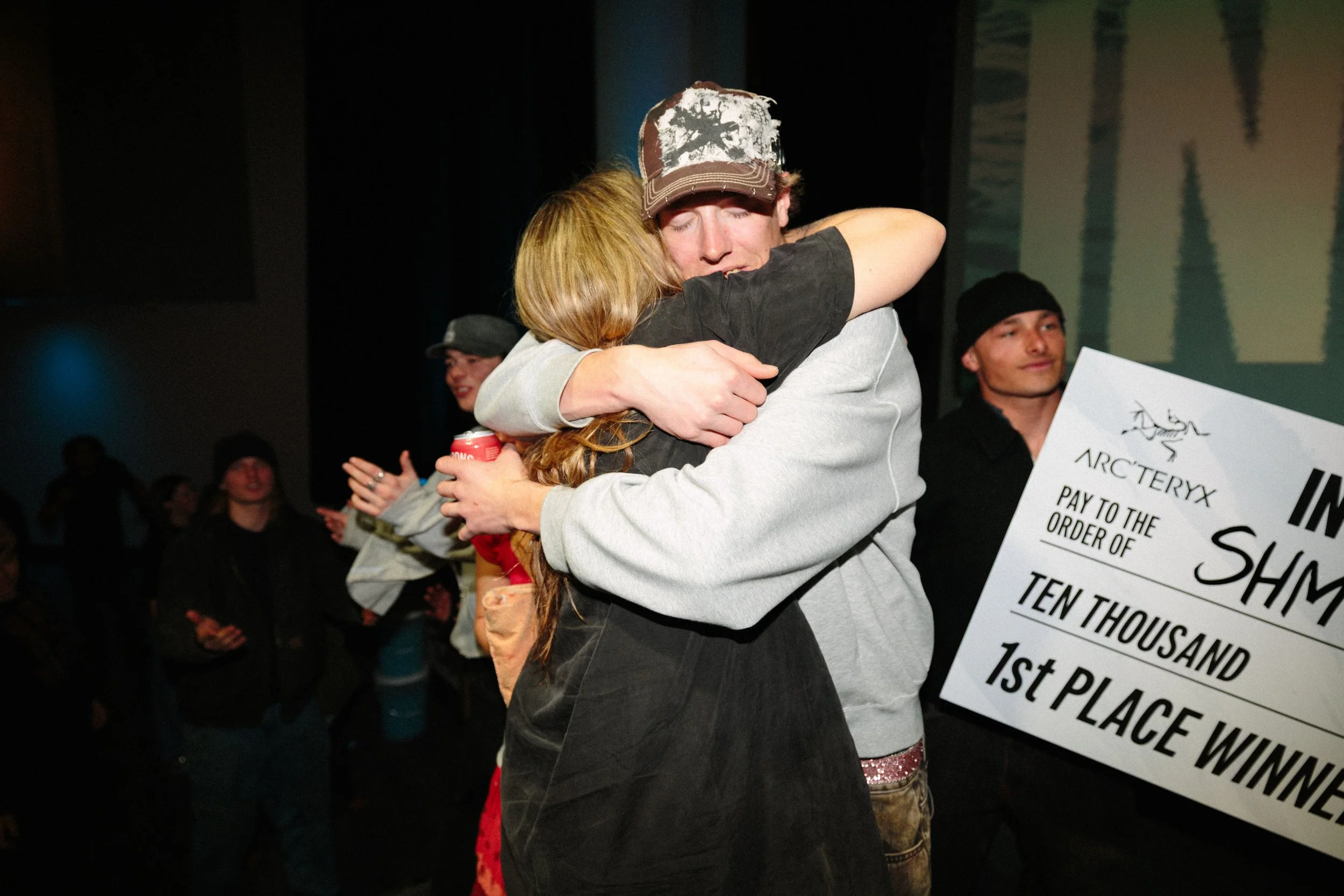 Two people hugging in a celebratory environment, with a crowd in the background, and a large check or award visible on the right side of the image.