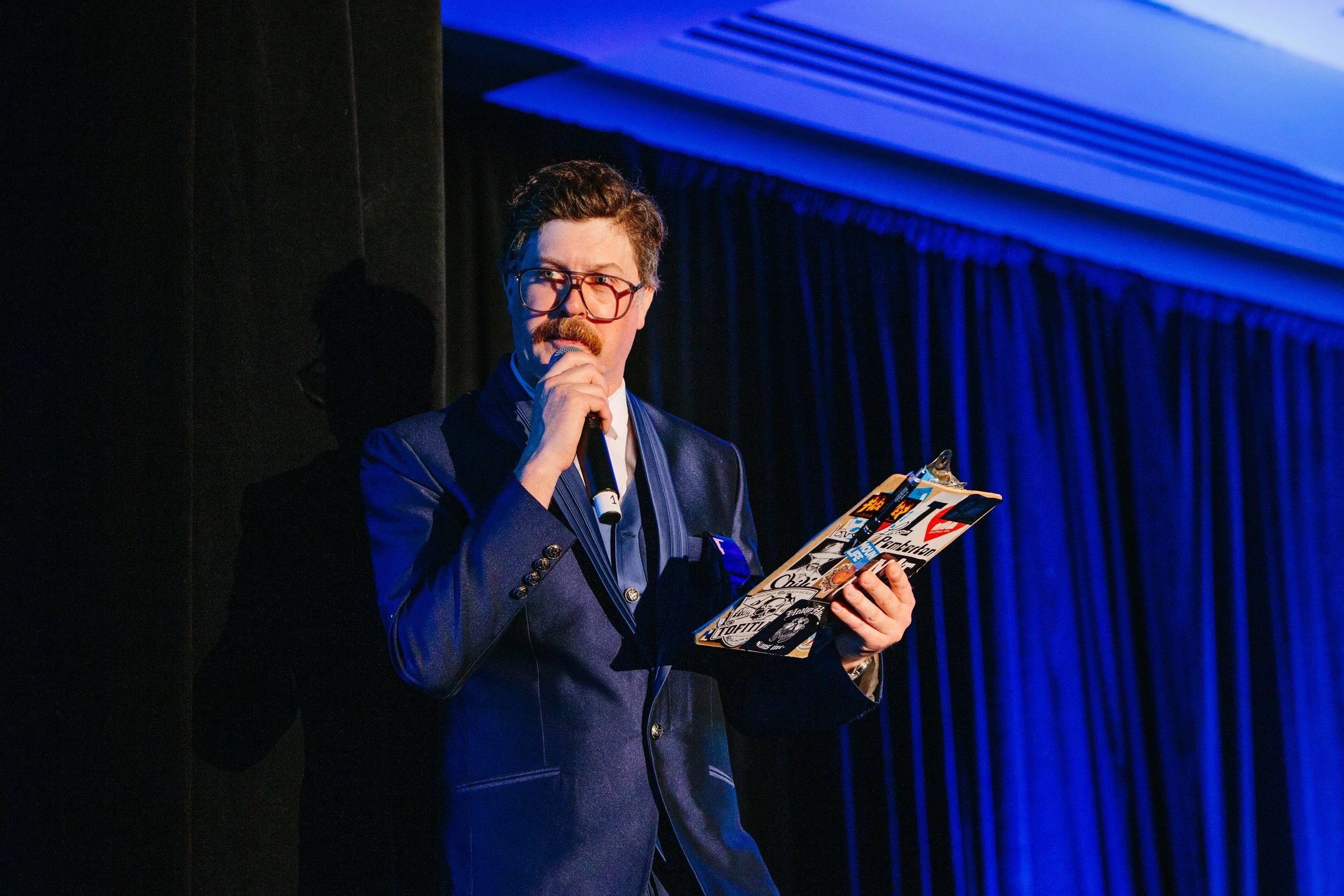 A man with glasses and a mustache, dressed in a blazer, is holding a microphone and reading a magazine while standing on a stage with blue curtains and stage lighting.