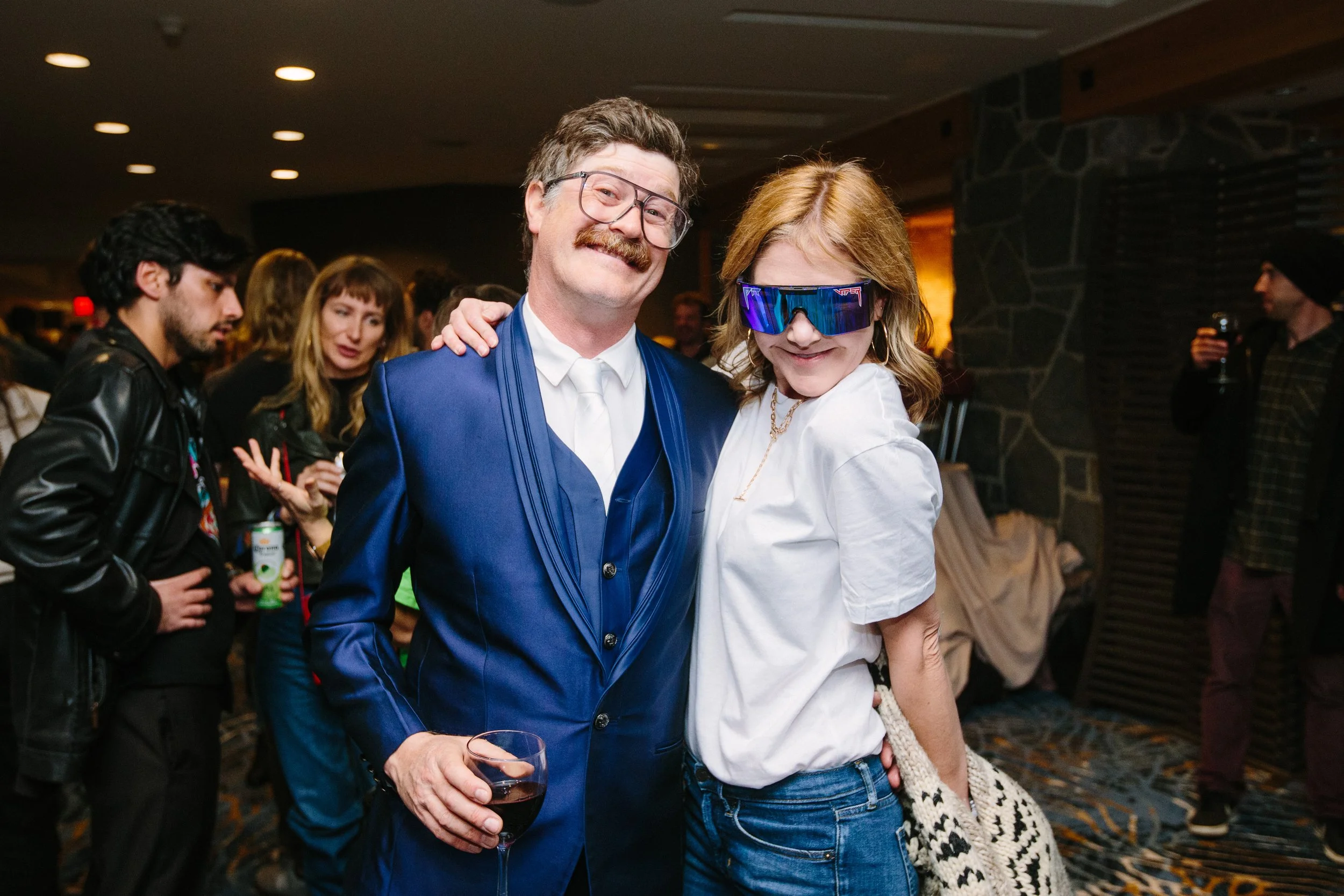 A man and a woman posing together at a social event, with the man holding a glass of red wine and the woman wearing large sunglasses, smiling at the camera. Other people are mingling in the background.
