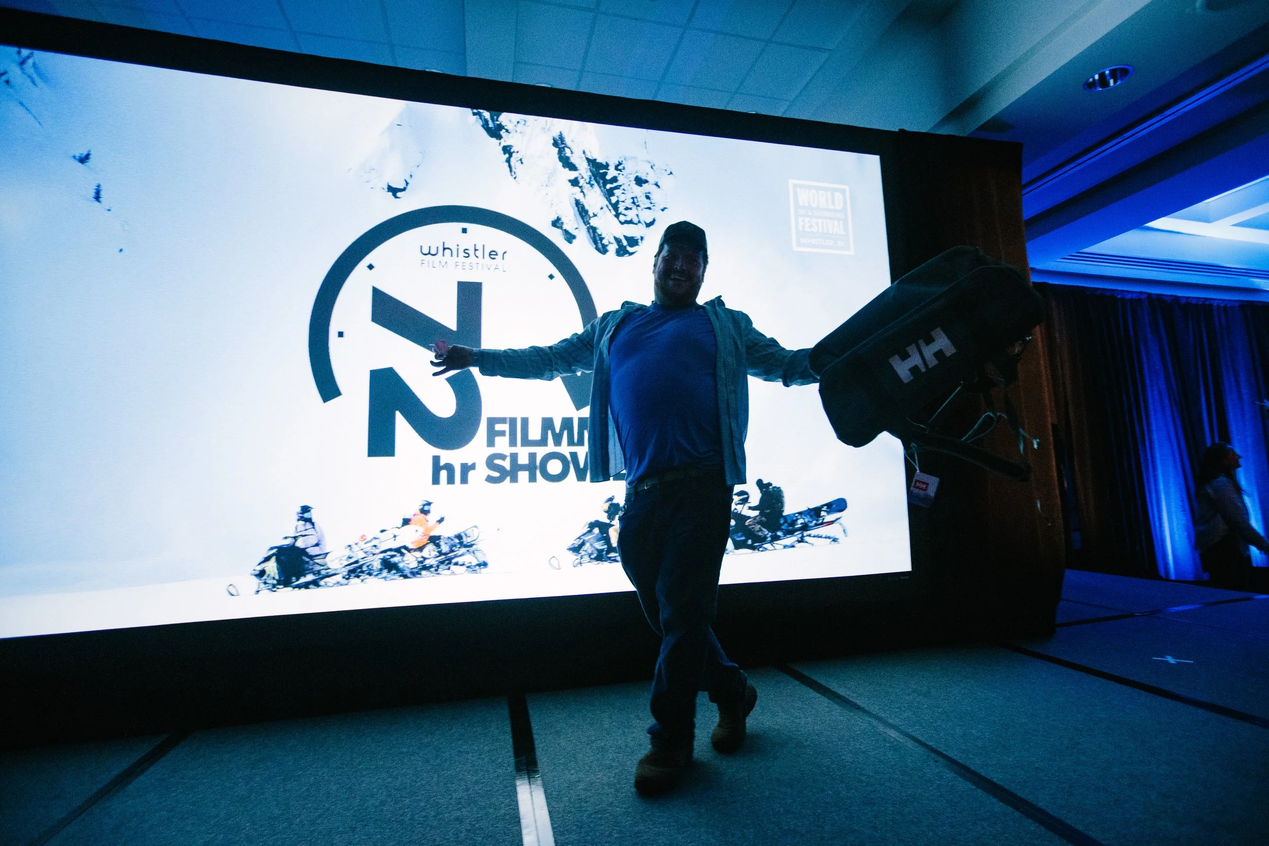 Person holding a camera bag standing in front of a large screen displaying the logo for the Whistler Film Festival and the text "22 hr FILM SHOW".