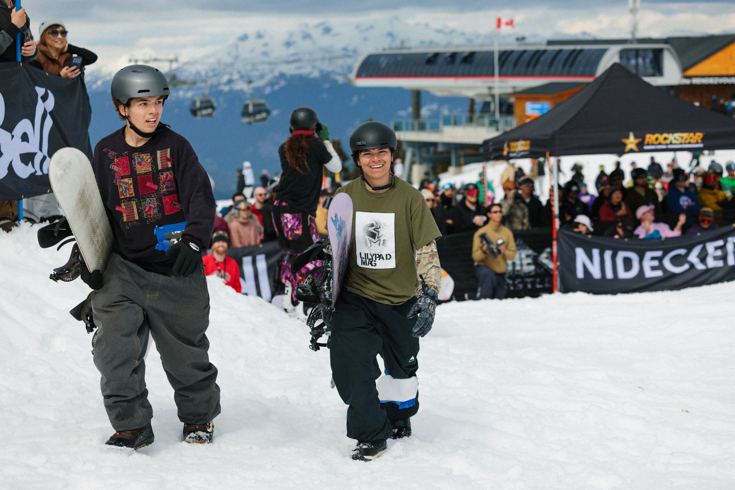 Two young snowboarders standing on snow at a ski resort, with a crowd of spectators behind them and a ski lift in the background.