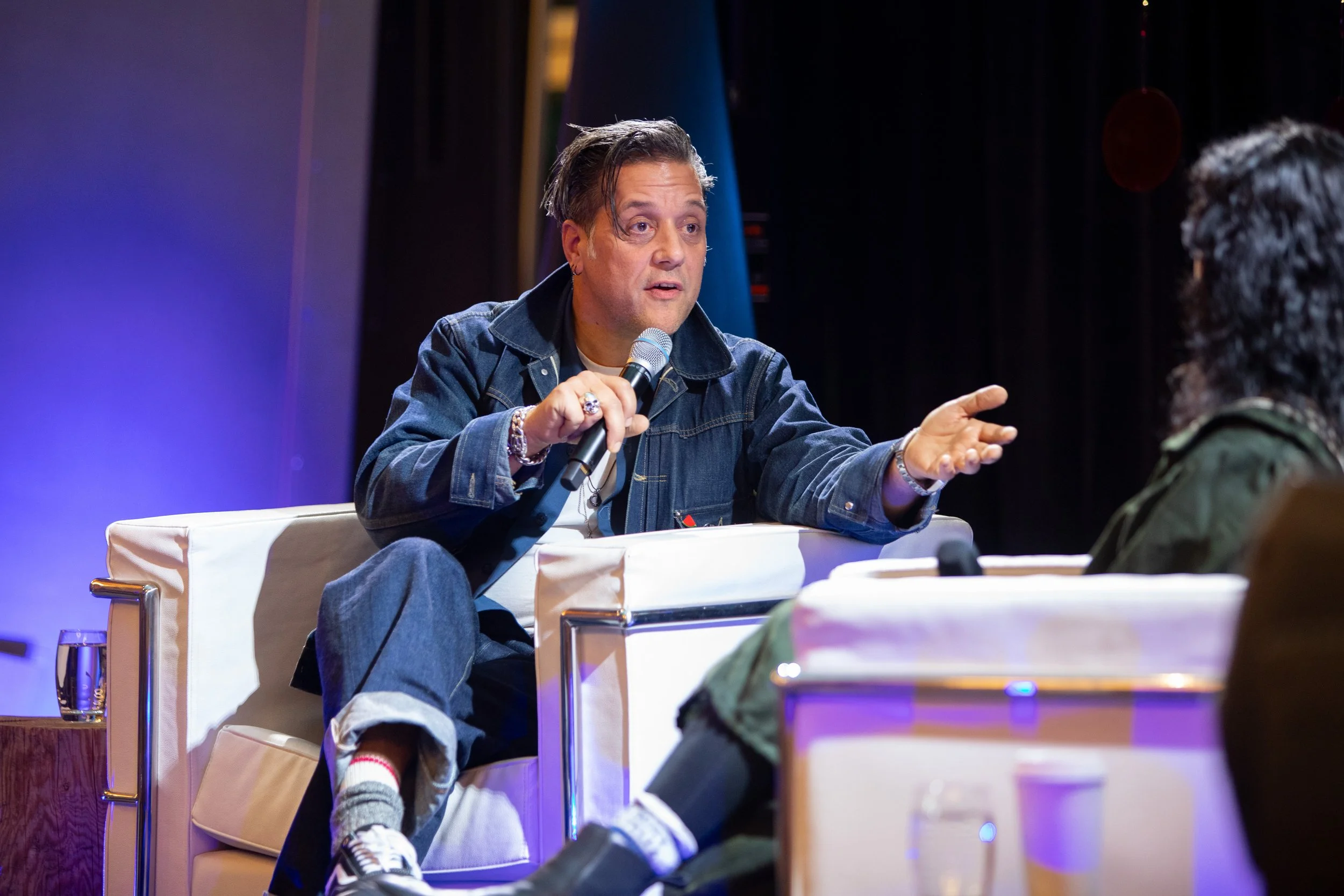 A man with dark hair, wearing a denim jacket, is sitting on a white armchair, holding a microphone, and talking to a woman with curly black hair, who is seated across from him in a dark setting with stage lighting.