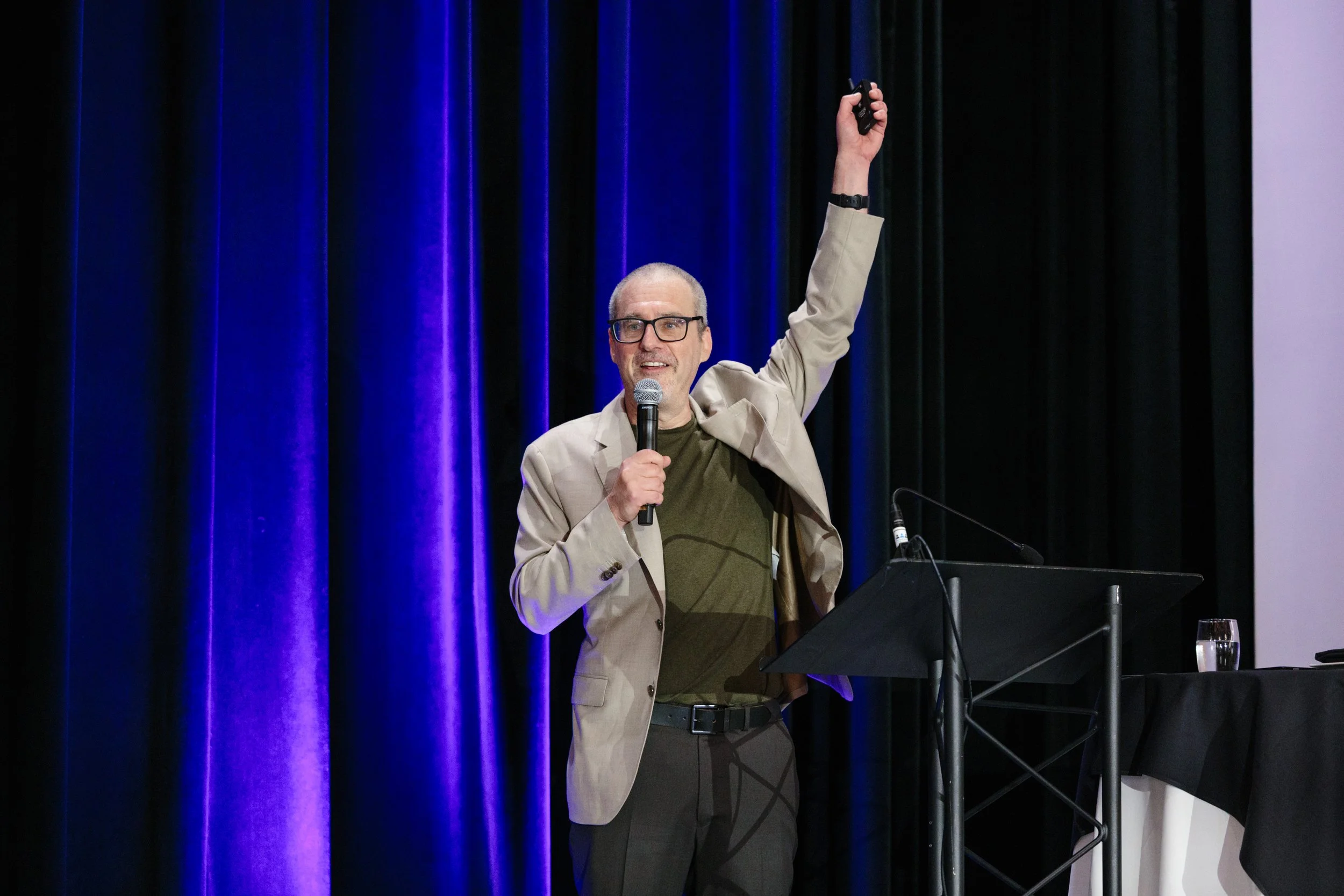 A man with glasses, wearing a beige blazer and dark pants, holding a microphone in one hand and raising his other hand, speaking to an audience on a stage with black and purple curtains.