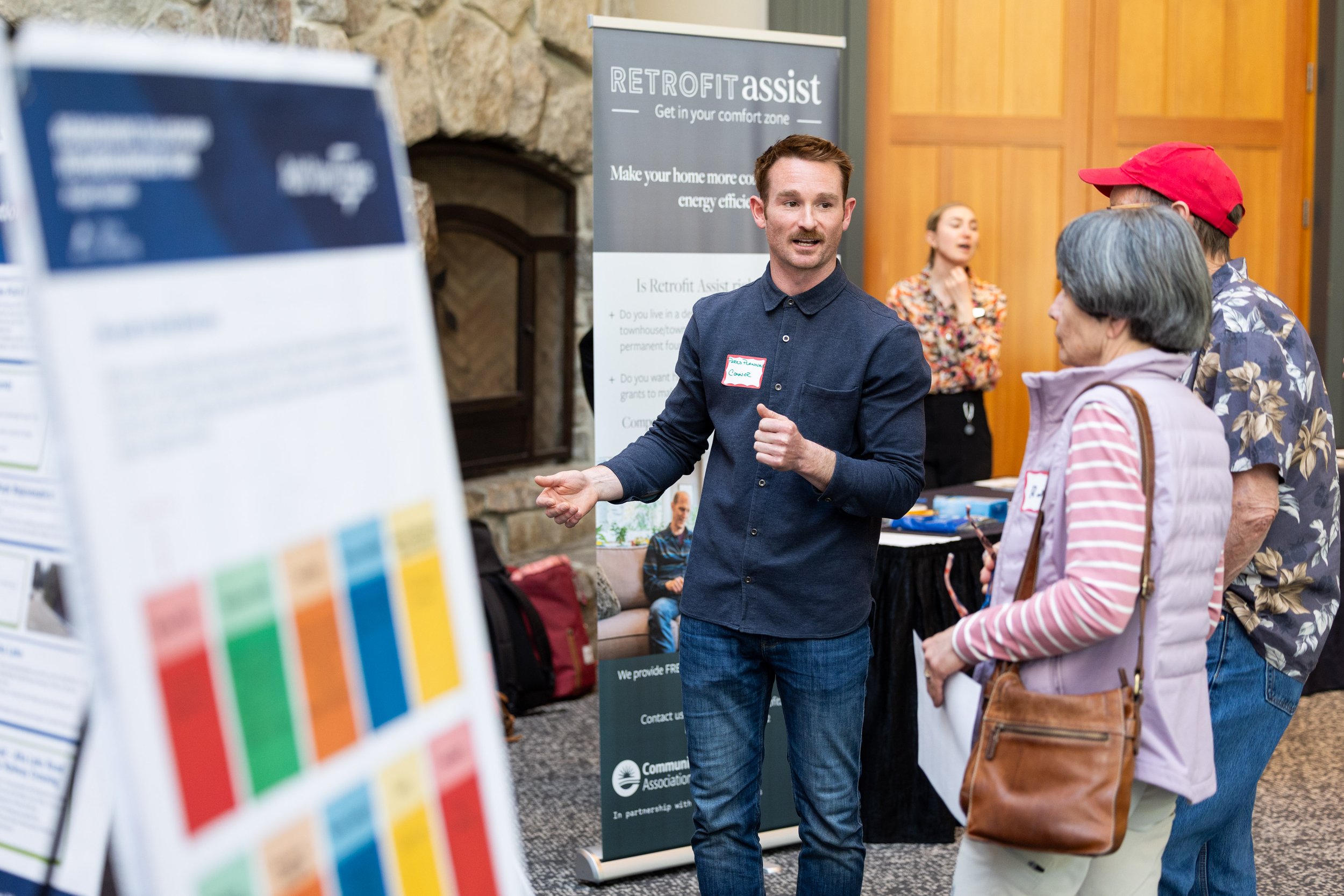 Man explaining event at a booth, with a RETROFIT assist banner and informational posters, engaging with three seniors in an indoor community event.