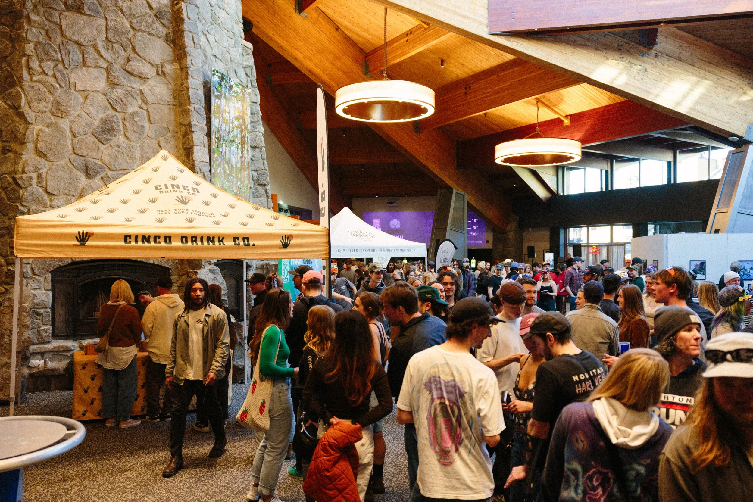 Crowd of people gathering indoors at an event with a stone fireplace, wooden ceiling, and hanging lights. Tents and displays are visible among the attendees.