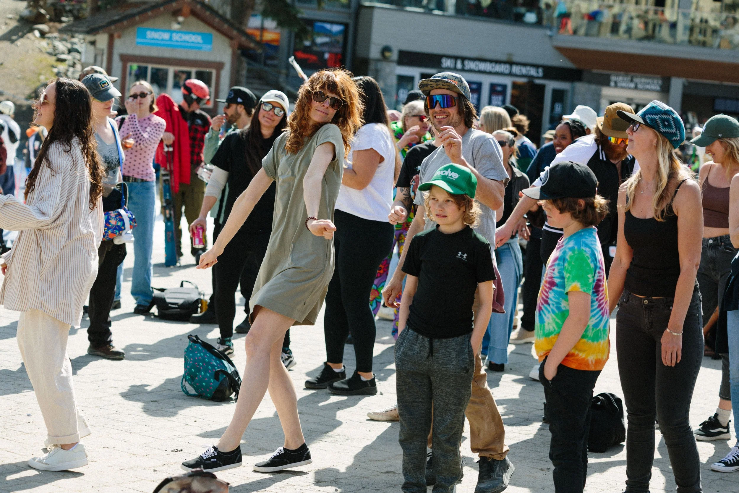 A group of people standing outdoors on a sunny day in front of shops. Some are smiling and wearing casual clothes, sunglasses, and hats.