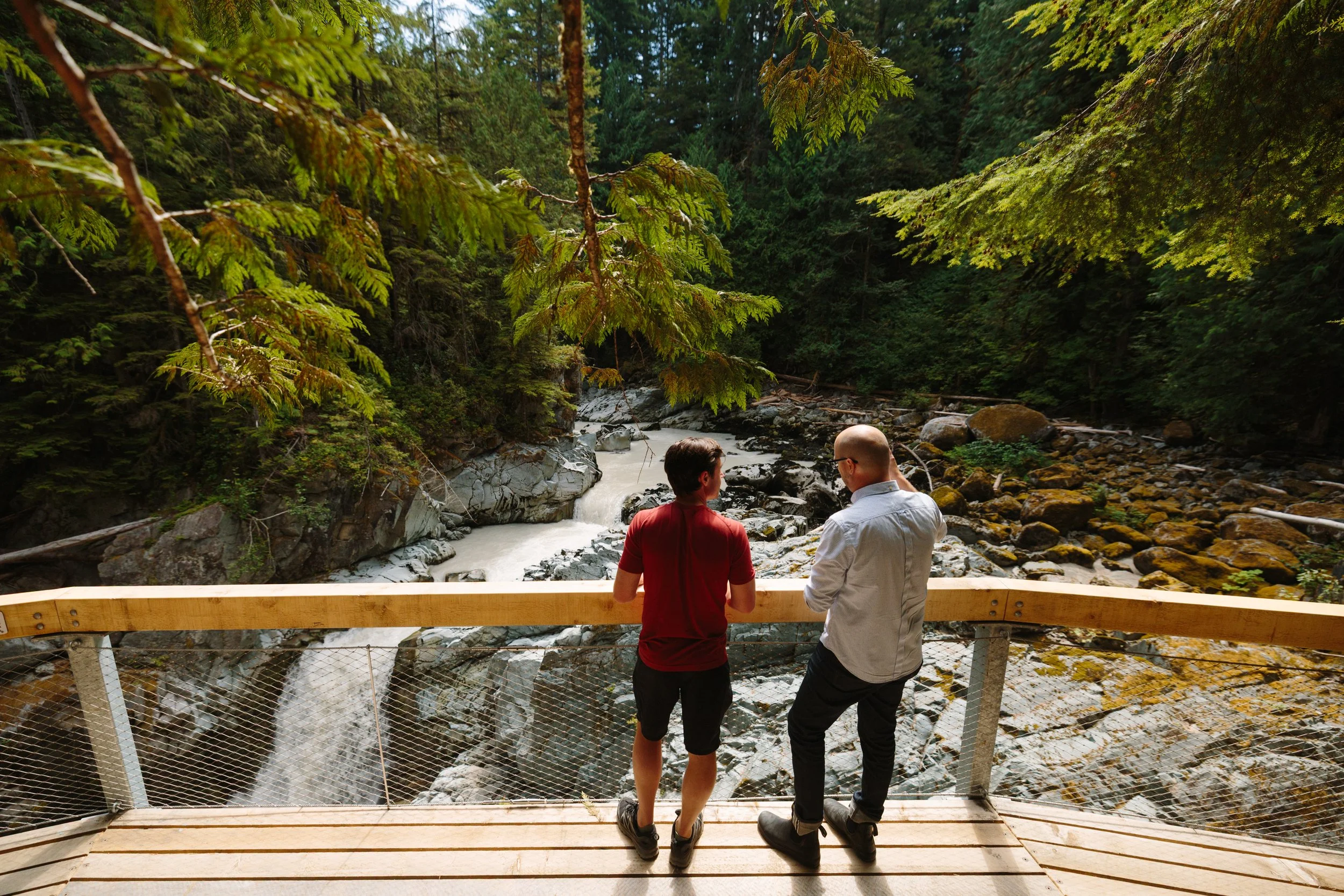Two men standing on a wooden viewing platform overlooking a rocky river in a forest.
