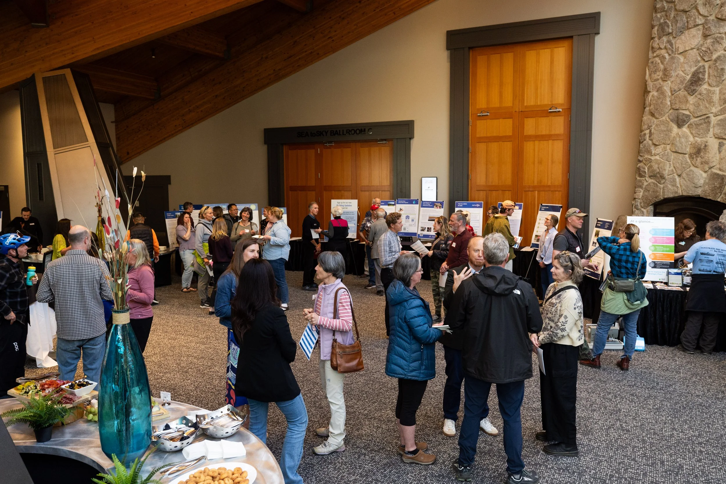 A busy conference or expo hall filled with attendees walking and talking, with various booths and posters on display, a table with food and a tall blue vase with decorative branches in the foreground.