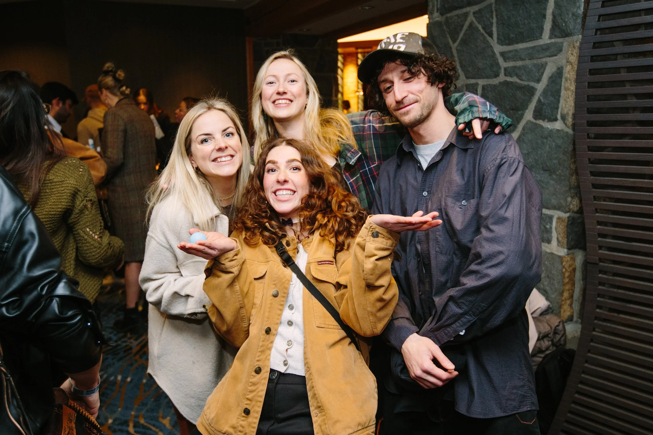 Group of four young people smiling at a social gathering, standing close together with one young woman holding a small object in her hand.