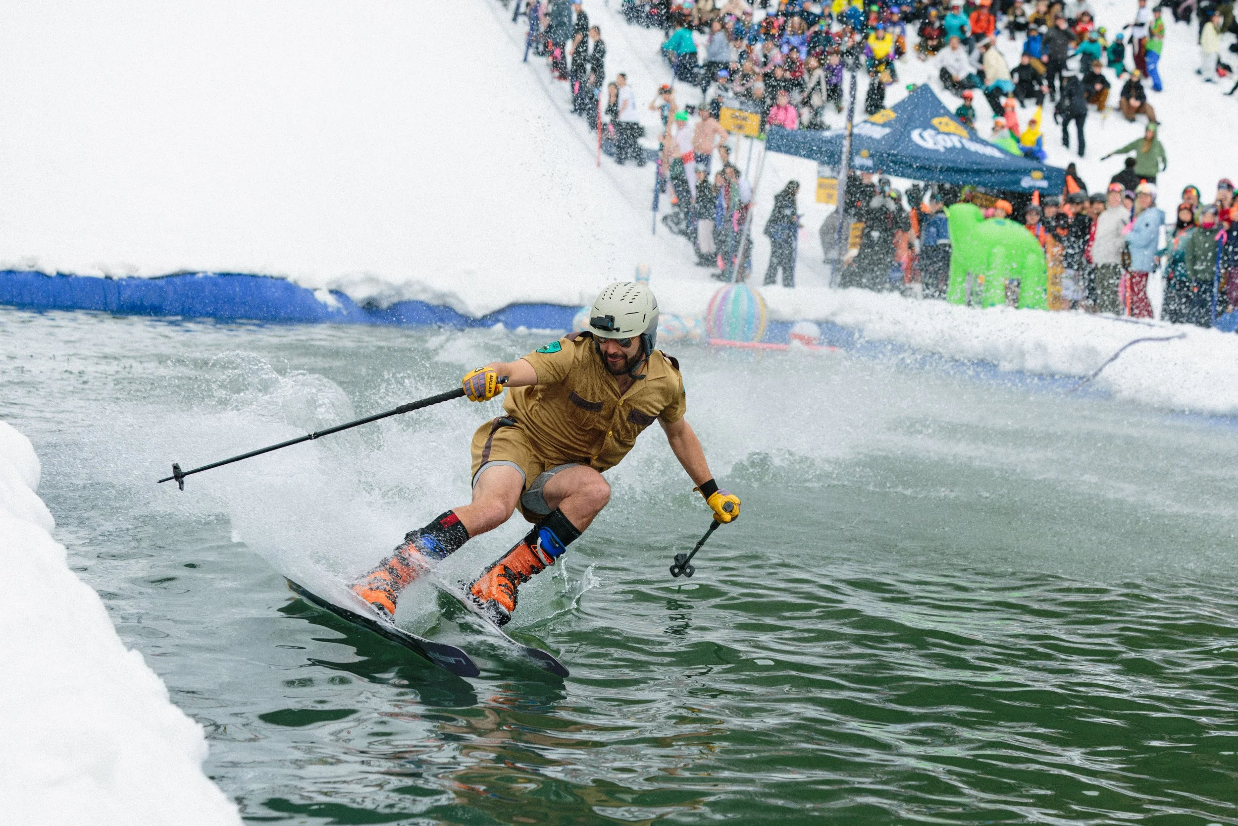 A man in a helmet and sunglasses skimboarding on water near the shoreline, while a large crowd of people gather on snowy land in the background.