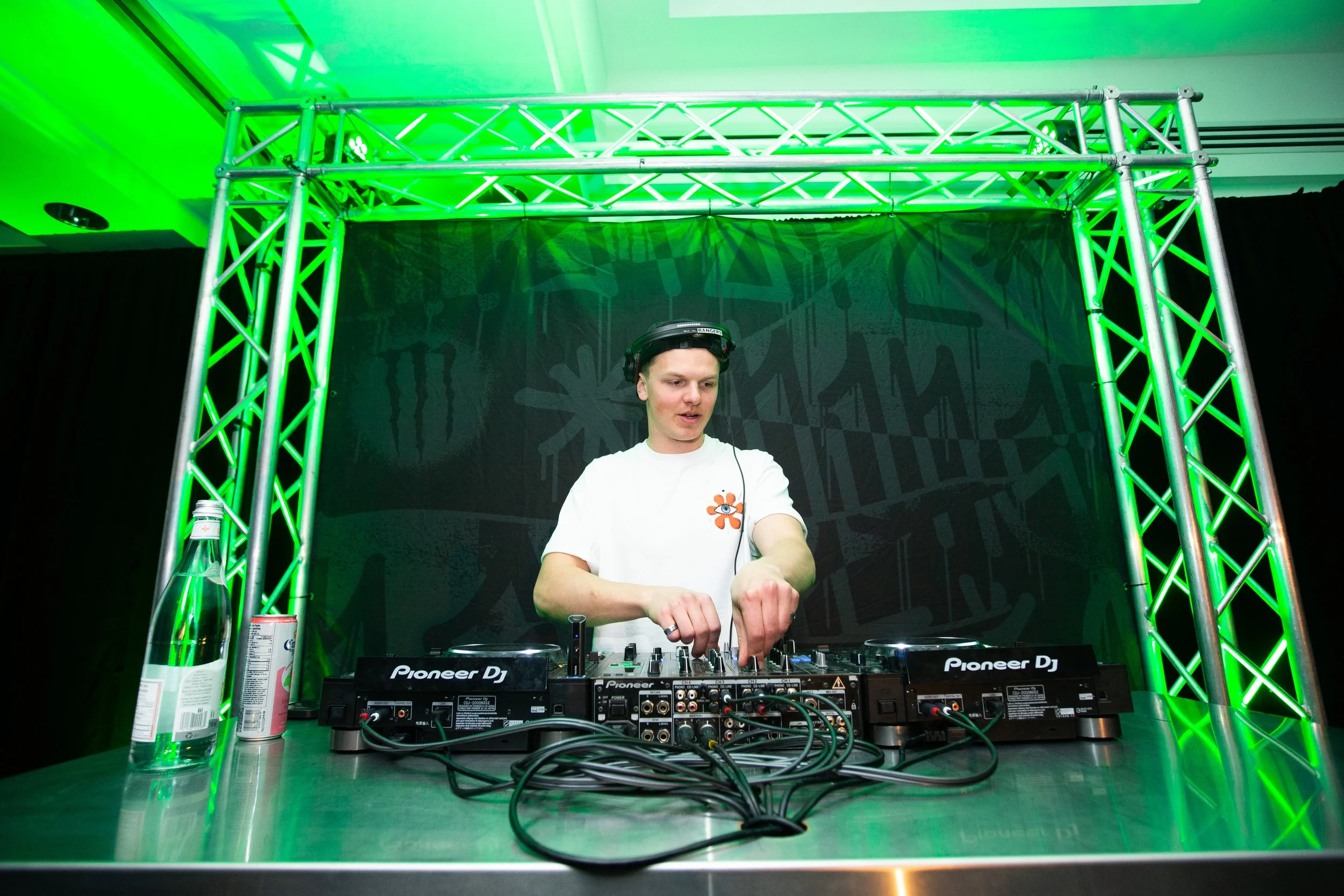 A DJ is performing at a nightclub or event, standing behind DJ equipment with a focused expression, surrounded by green lighting and a truss structure.