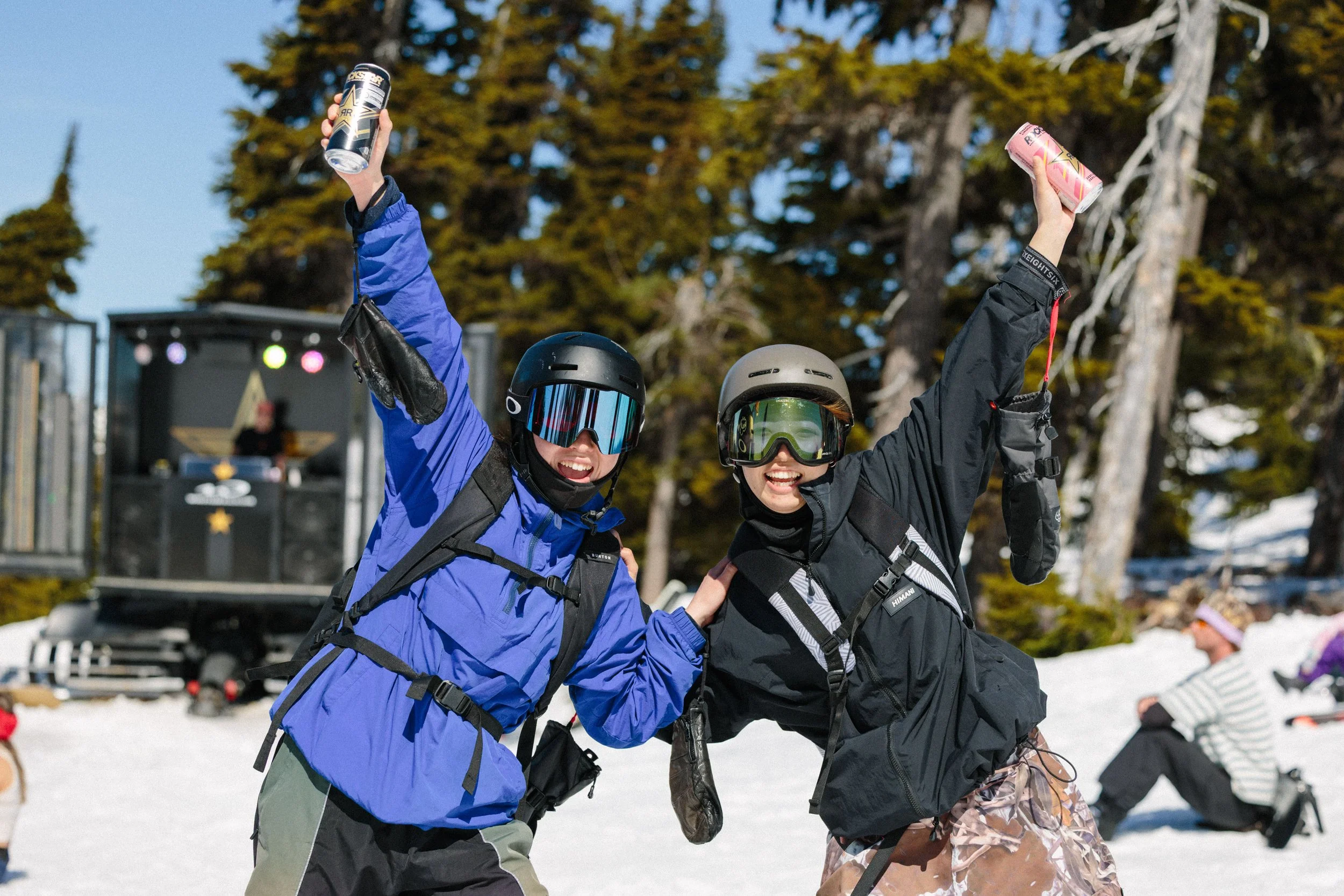 Two people wearing ski gear, helmets, and goggles, celebrating outdoors on snow; they are holding up cans of drinks and smiling.