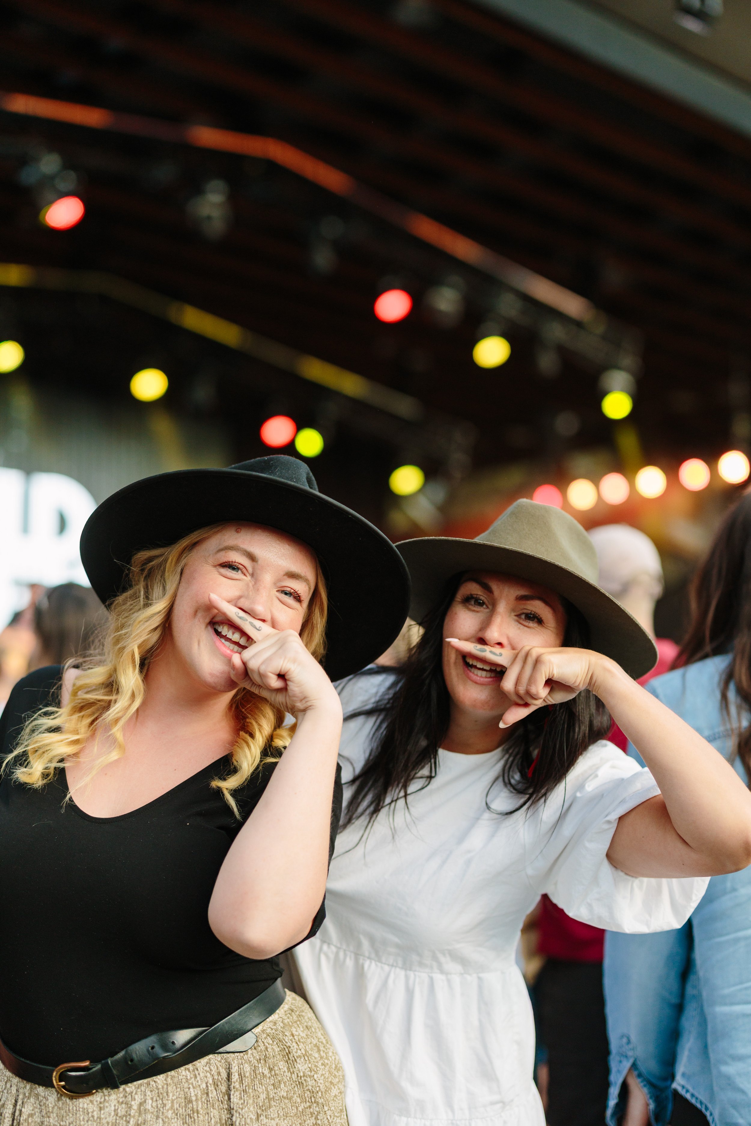 Two women smiling and wearing large hats, posing at a lively outdoor event or concert with colorful lights overhead.