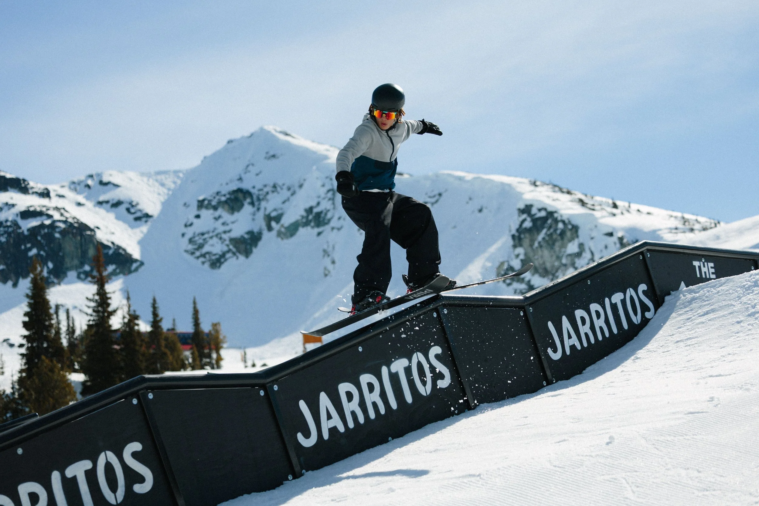 Person wearing a helmet and sunglasses snowboarding on a rail in a snowy mountain landscape.