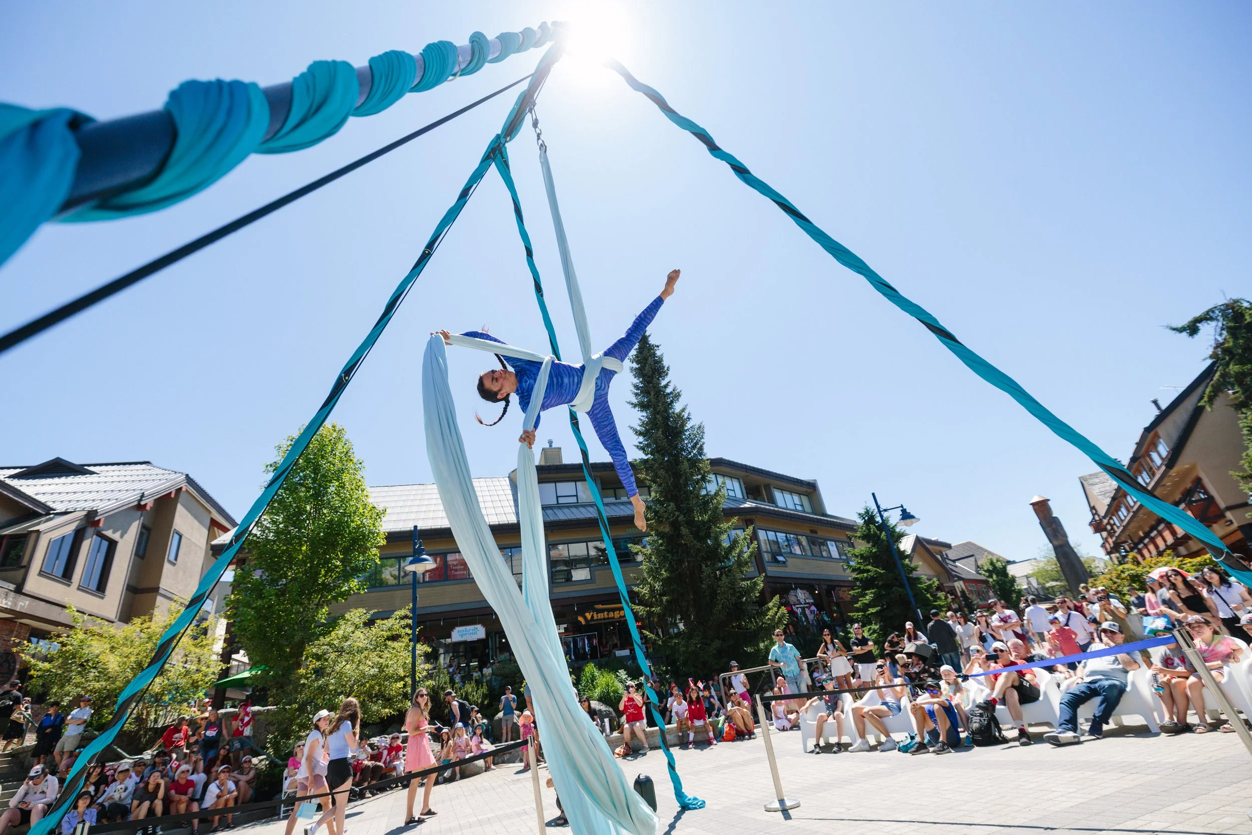 Circus performer demonstrating aerial silk act on a sunny day in a busy town square with spectators watching.