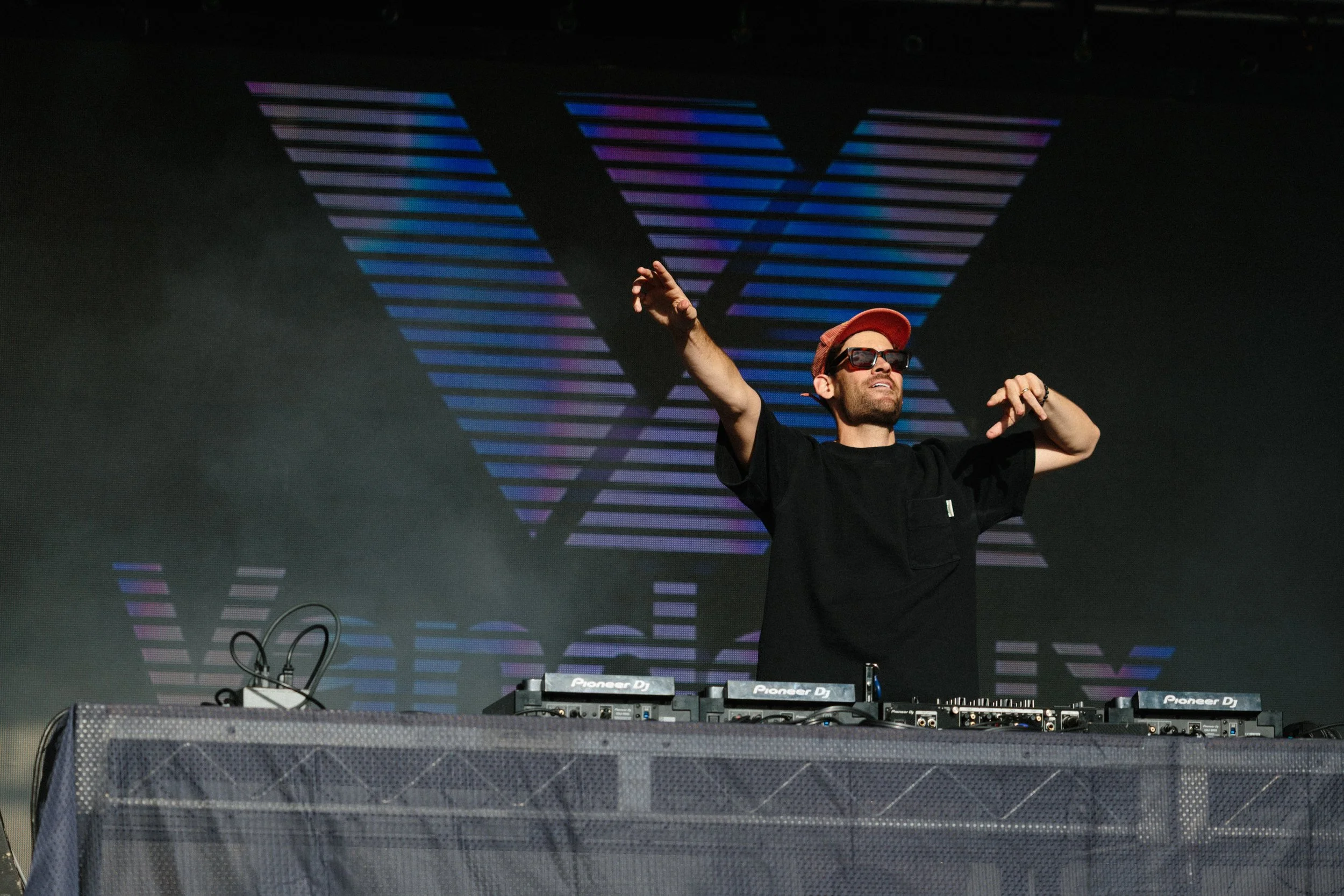 A man wearing sunglasses and a red baseball cap with a black t-shirt is DJing at a music event, standing behind a Pioneer DJ equipment setup. He is raising one arm and appears to be enjoying the music, with a large digital display with colorful strip