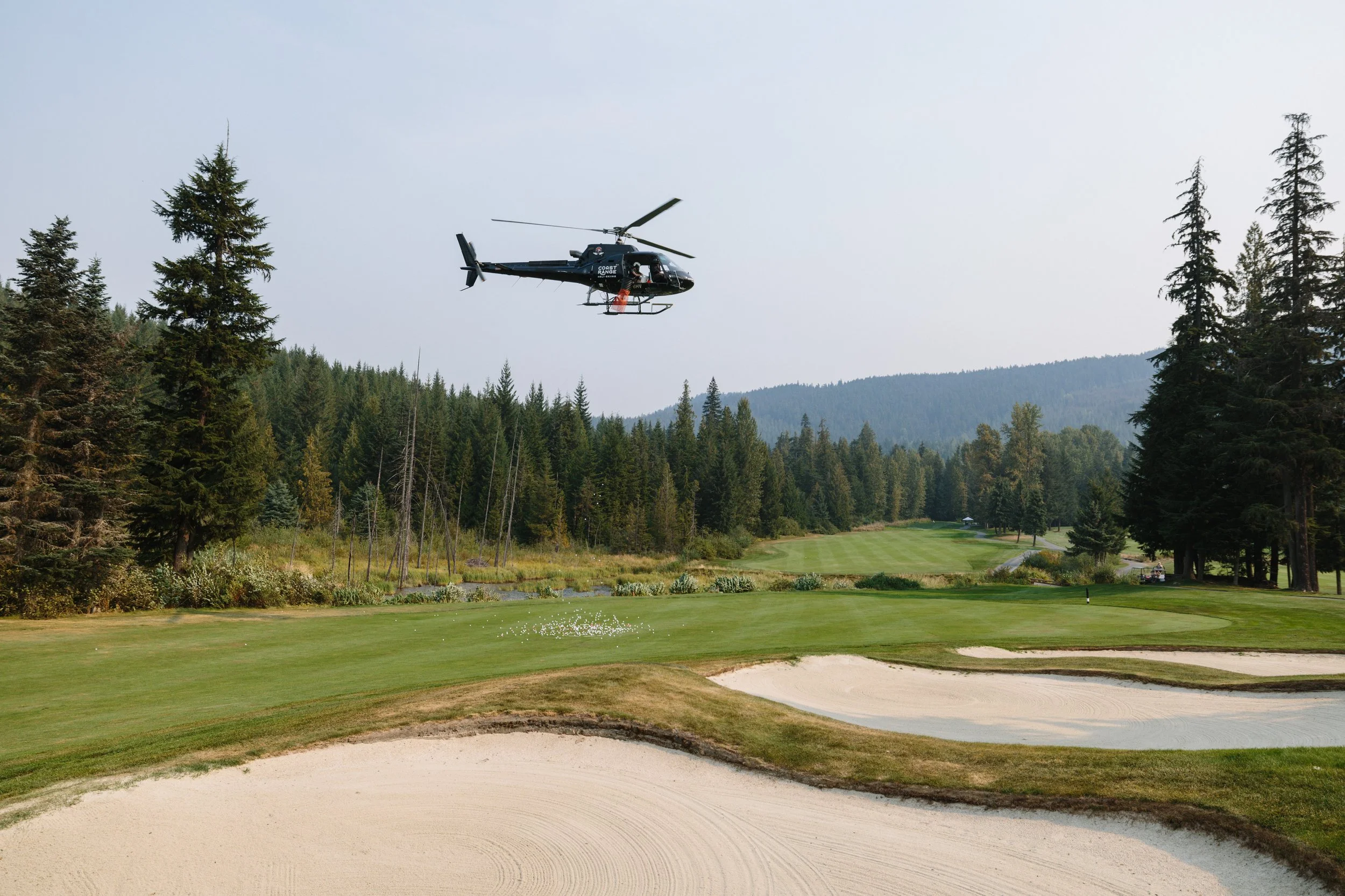 A helicopter flying over a golf course with sand traps and green fairways surrounded by trees and mountains.