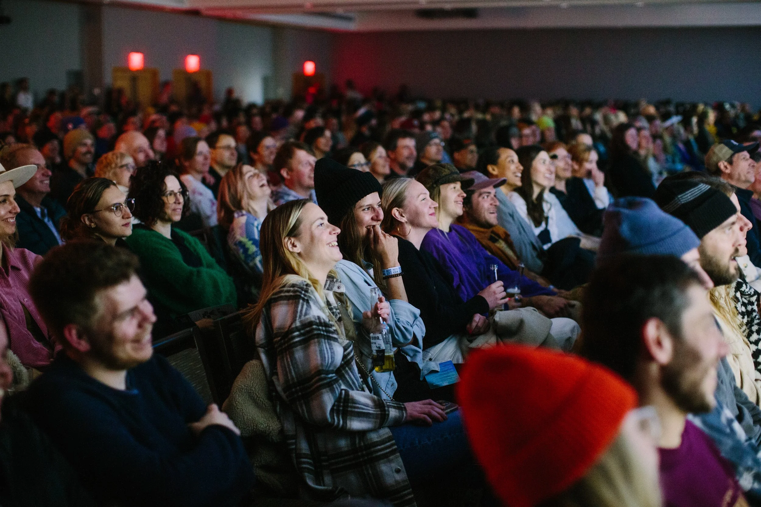 Audience sitting in a dark theater or conference hall, watching a performance or presentation, with focused and amused expressions.