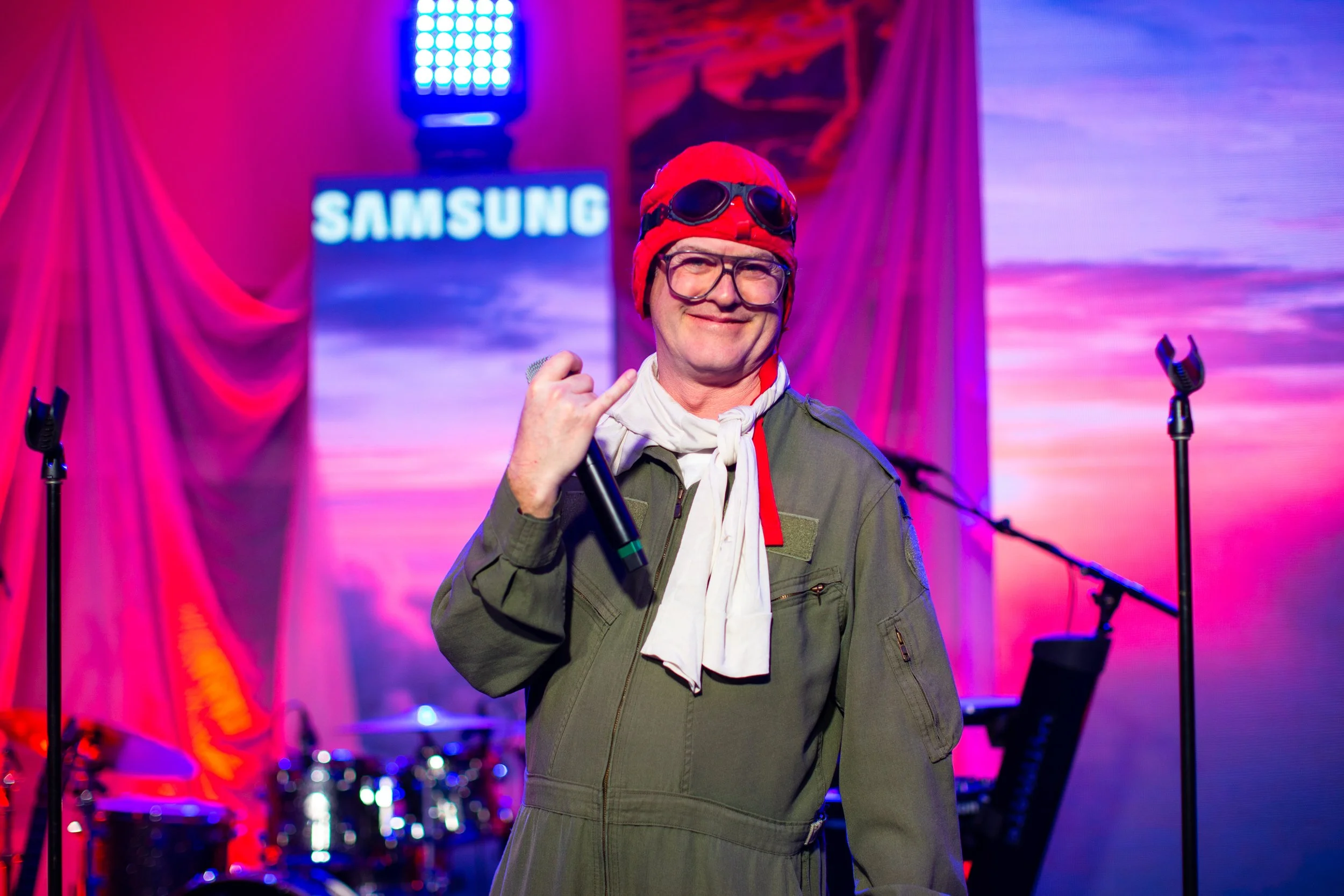 Man dressed in a vintage pilot costume, holding a microphone, standing on a stage with pink and purple lighting, a drum set and a Samsung sign in the background.