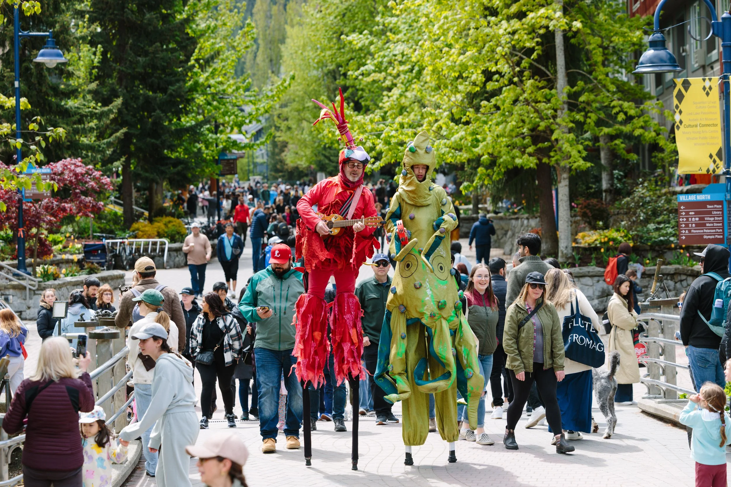 Street performers on stilts dressed in colorful costumes entertaining a crowd in a park during springtime. The scene is lively with many spectators, some taking photos and some walking.