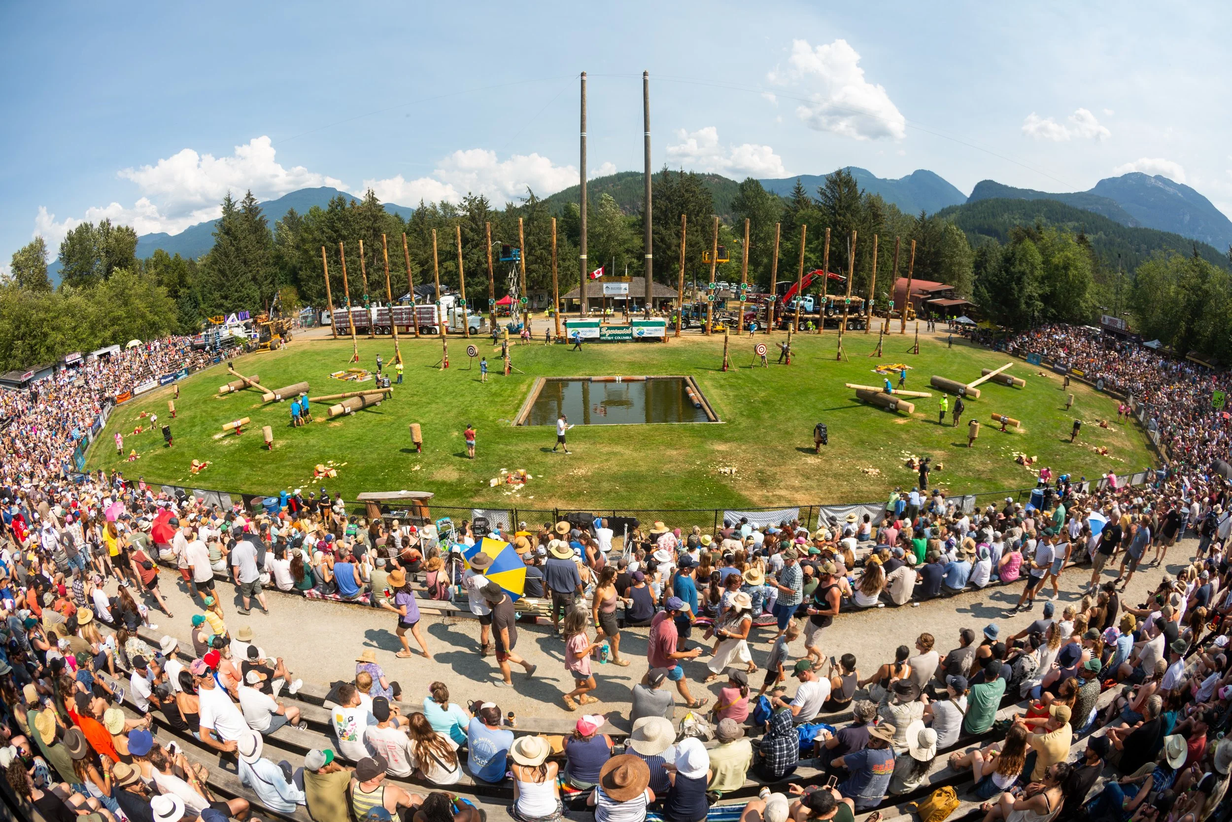 A large crowd watching a lumberjack competition in an outdoor arena with mountains in the background.