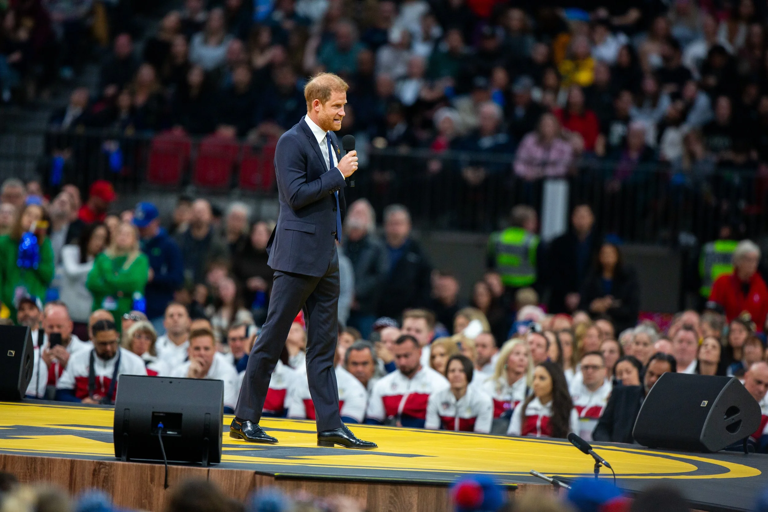 Prince Harry speaking into a microphone on stage at a public event with a seated crowd in the background.